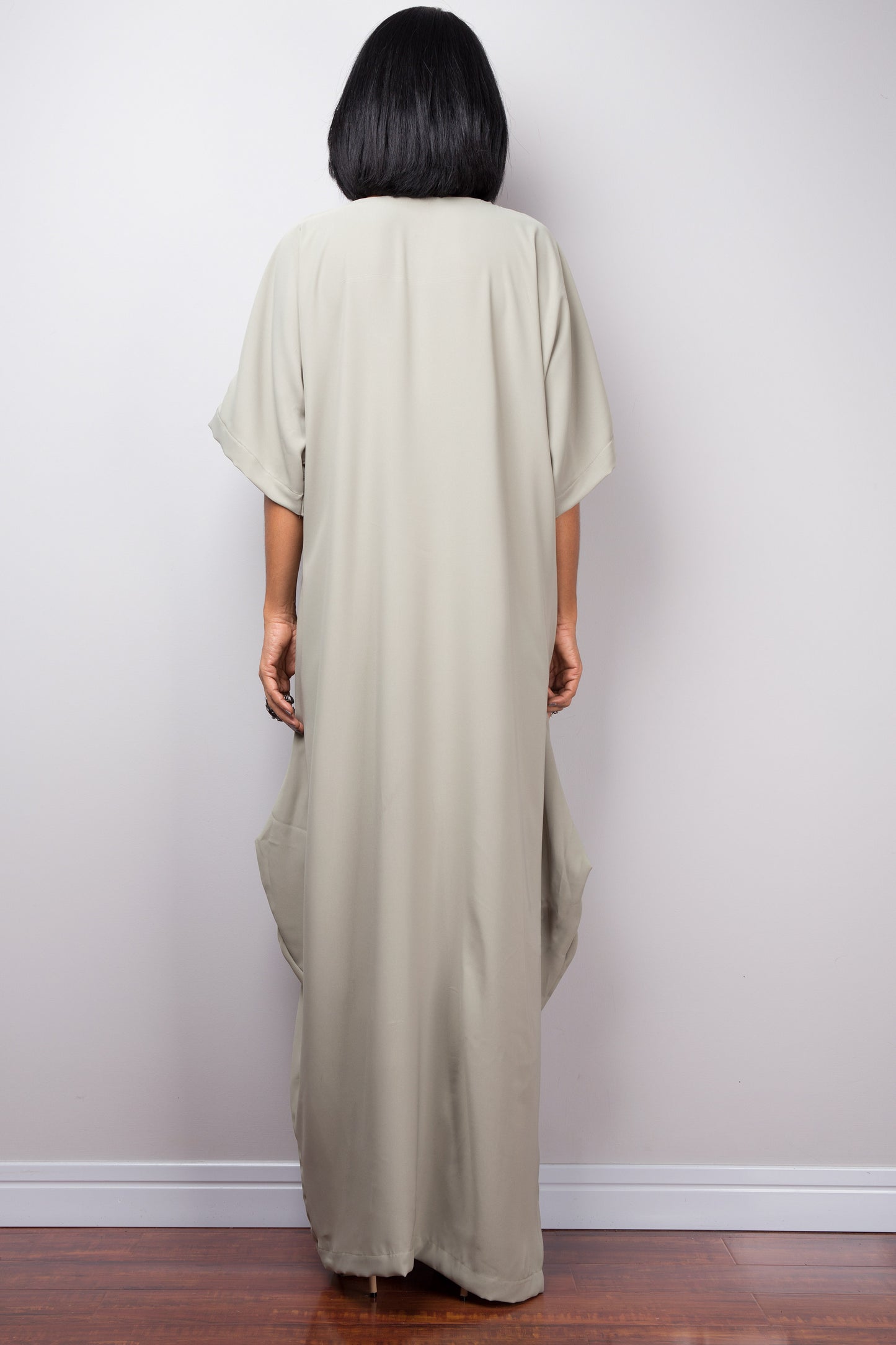 
Sage green kaftan maxi dress by Nuichan, worn by a woman with short black hair, standing gracefully against a gray wall and wooden floor, back view.

