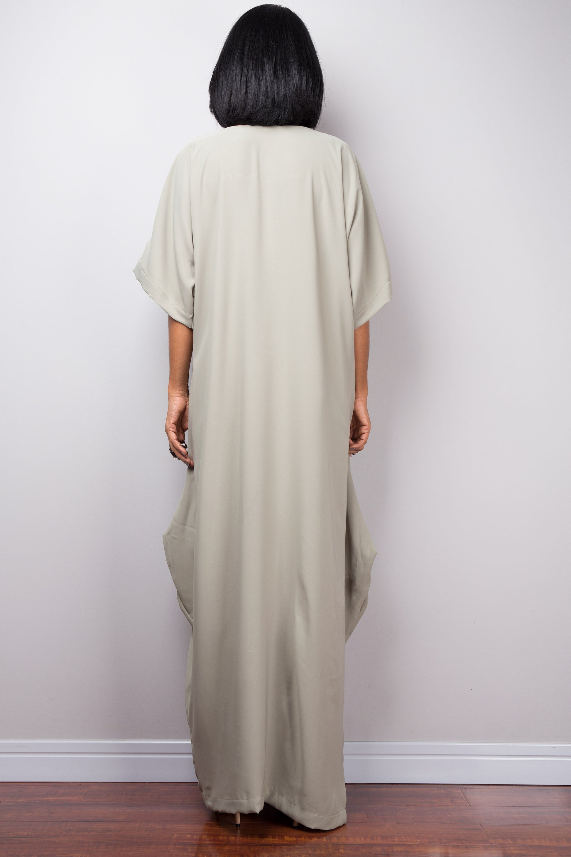 
Sage green kaftan maxi dress by Nuichan, worn by a woman with short black hair, standing gracefully against a gray wall and wooden floor, back view.

