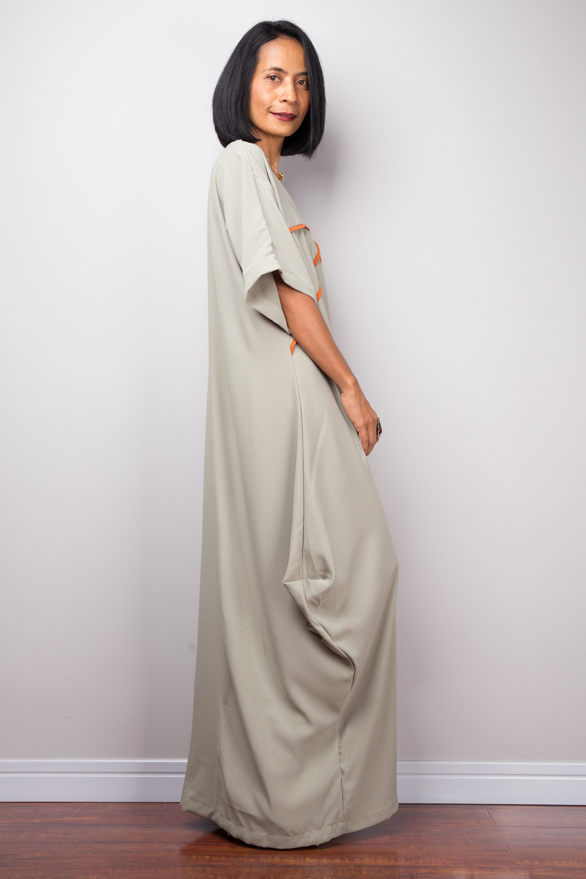 A floor-length sage kaftan dress from Nuichan, with white crisscross chest detail, worn by a woman standing sideways, set against a gray wall and wooden floor.

