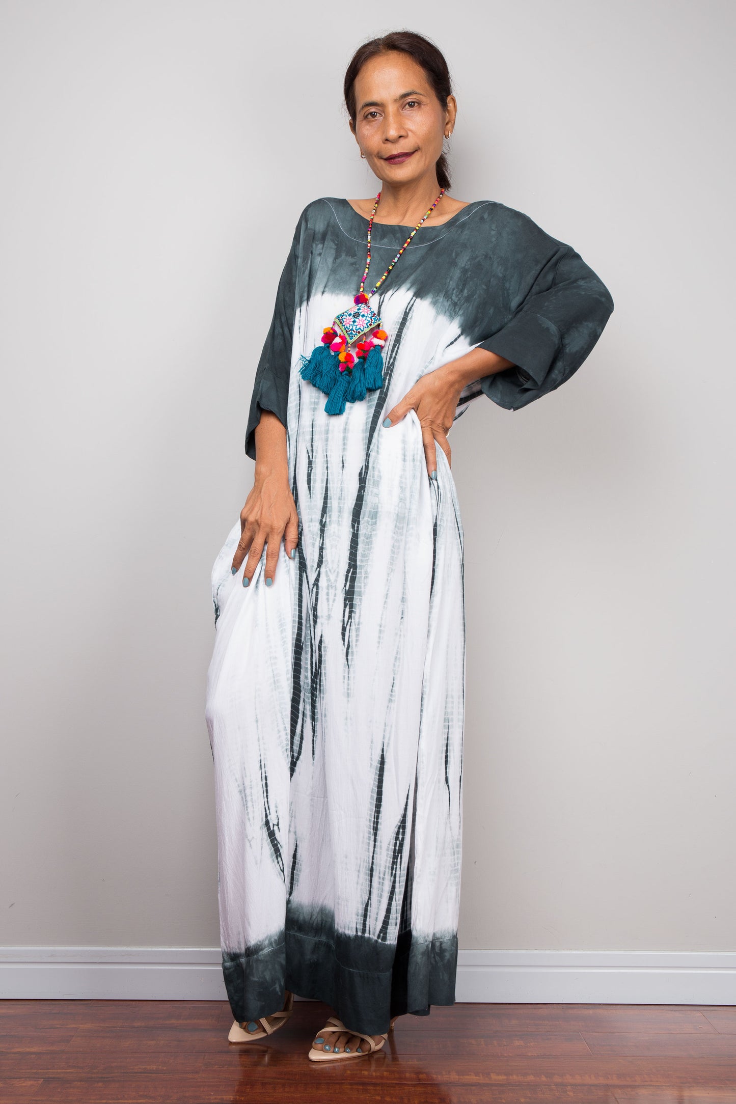 Woman wearing a long, dark gray and white shibori tie dye dress with a colorful pendant against a plain background