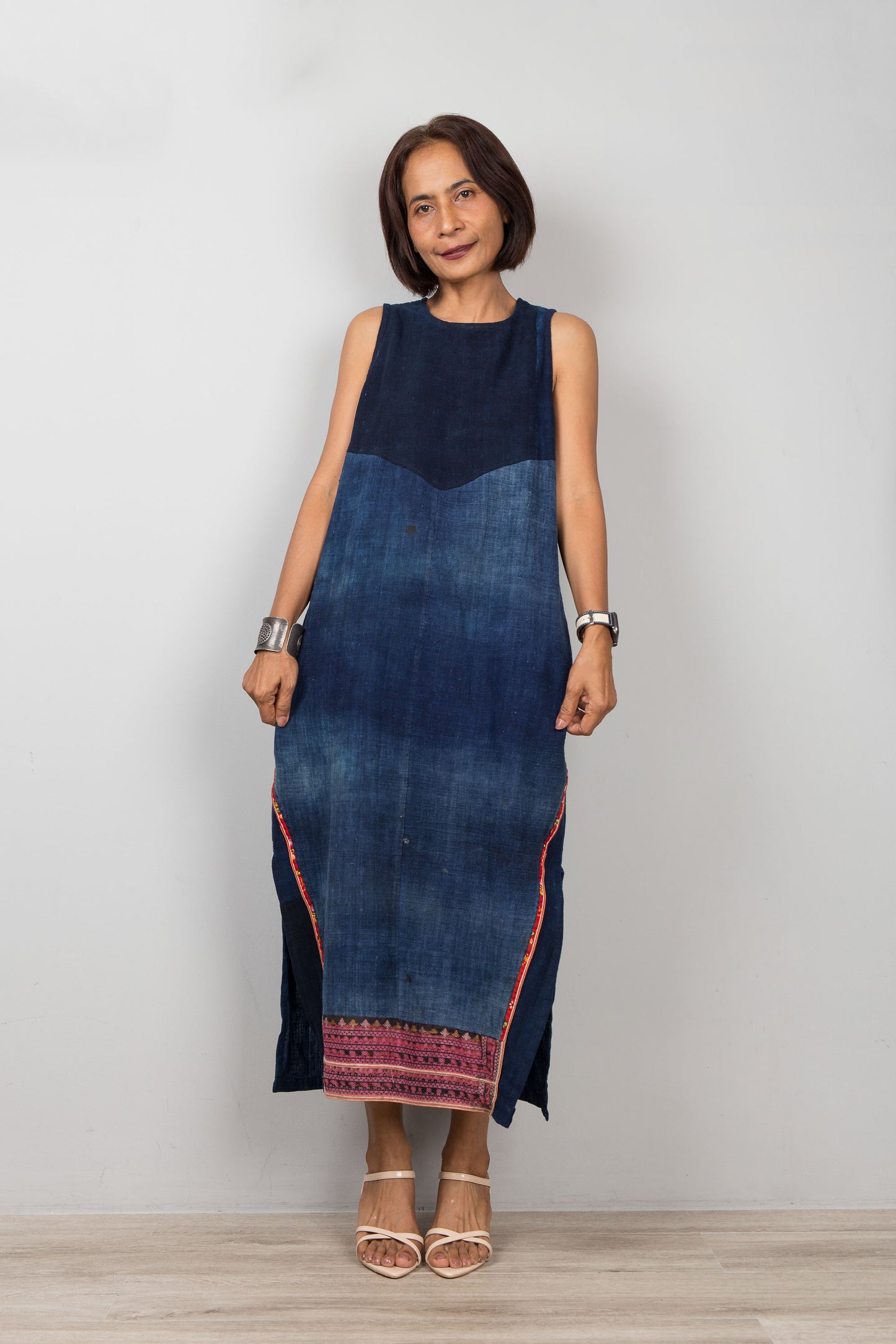 Woman wearing a blue, sleeveless, cotton dress with embroidery, pockets and a slit on a plain background