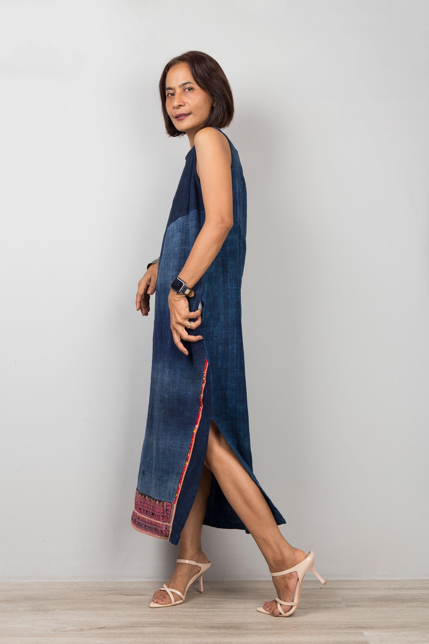 Woman wearing a blue sleeveless petite dress with embroidery, pockets and a slit on a plain background