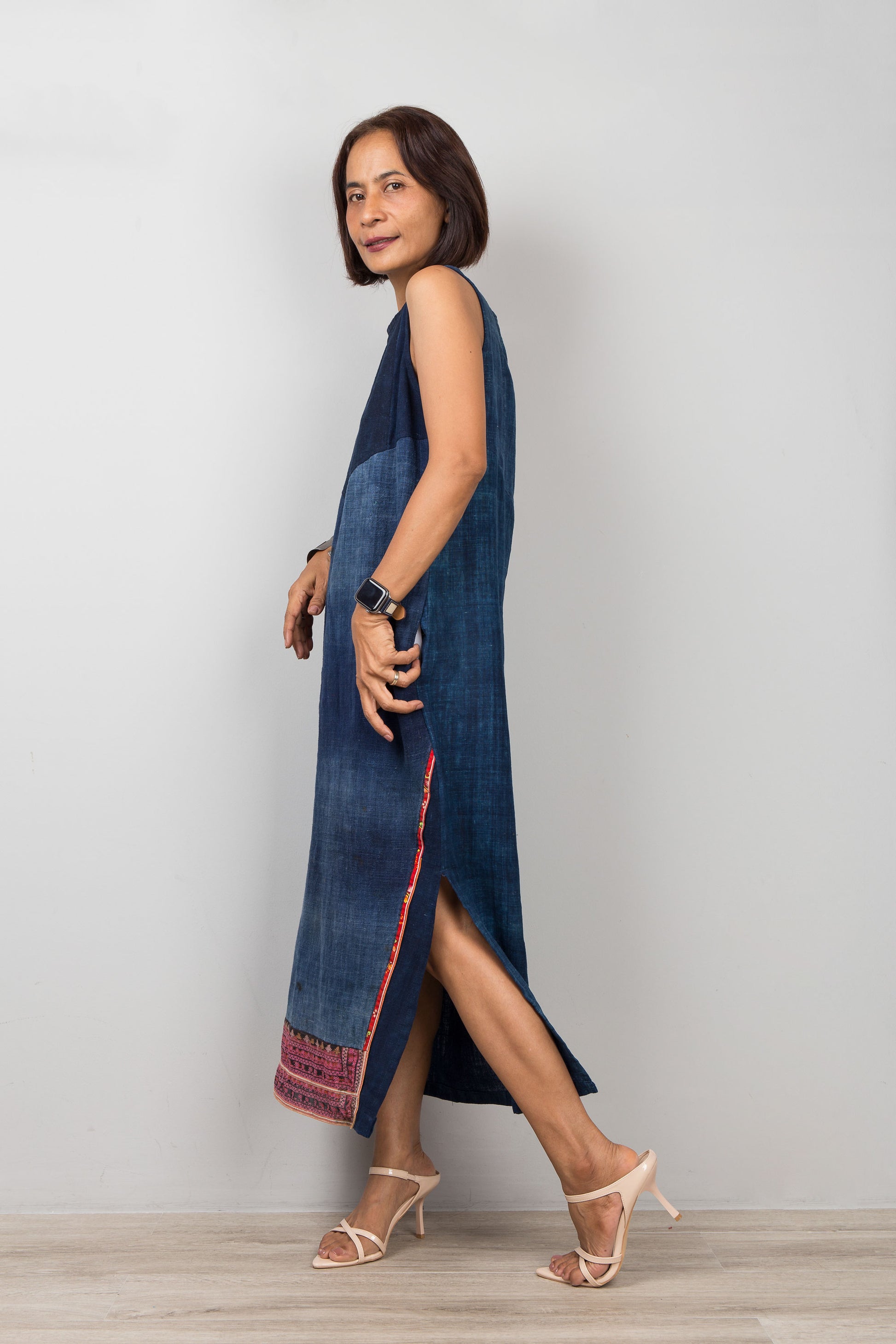 Woman wearing a blue sleeveless petite dress with embroidery, pockets and a slit on a plain background