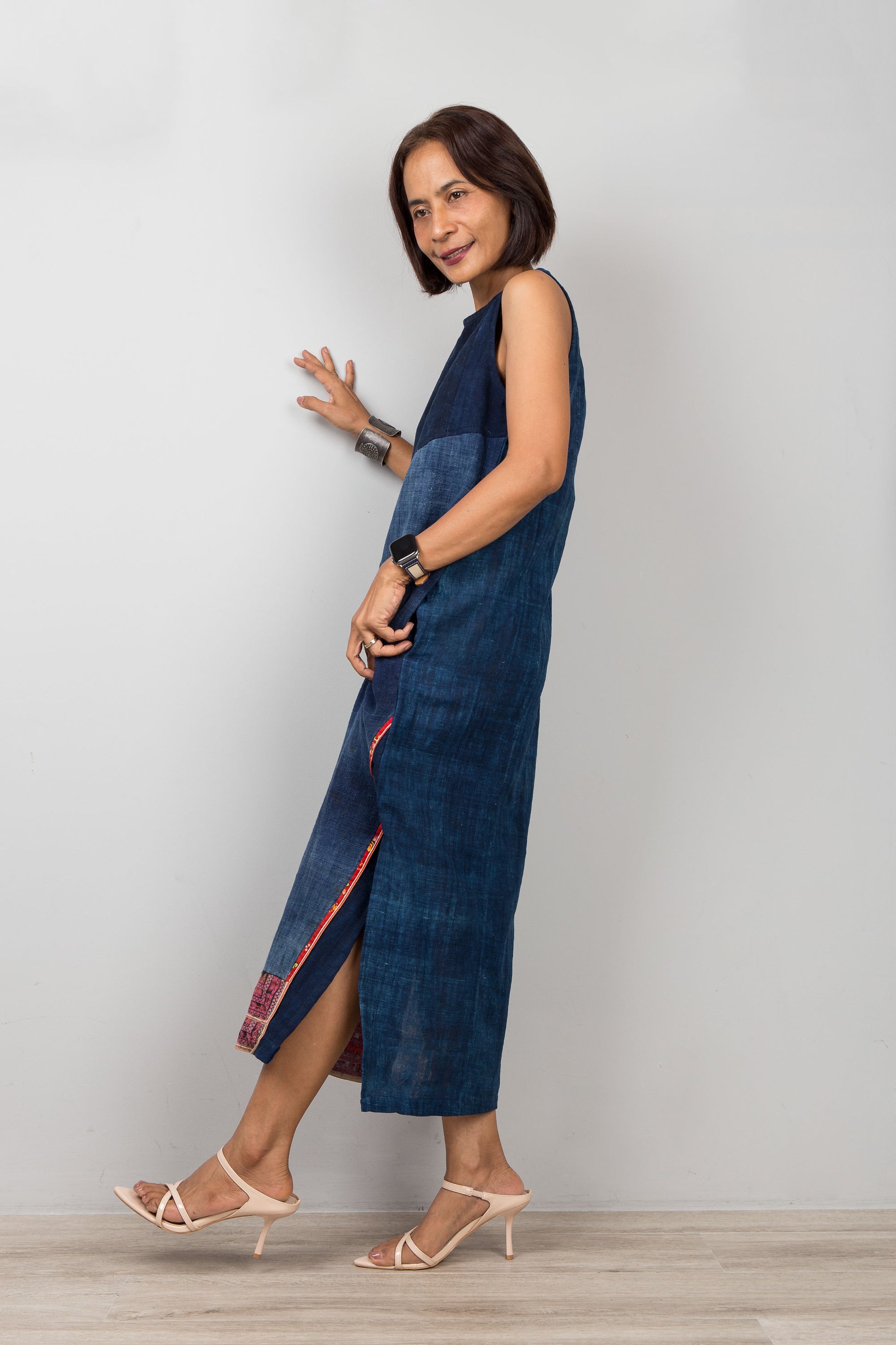 Woman wearing a blue cotton midi dress with embroidery, pockets and a slit on a plain background