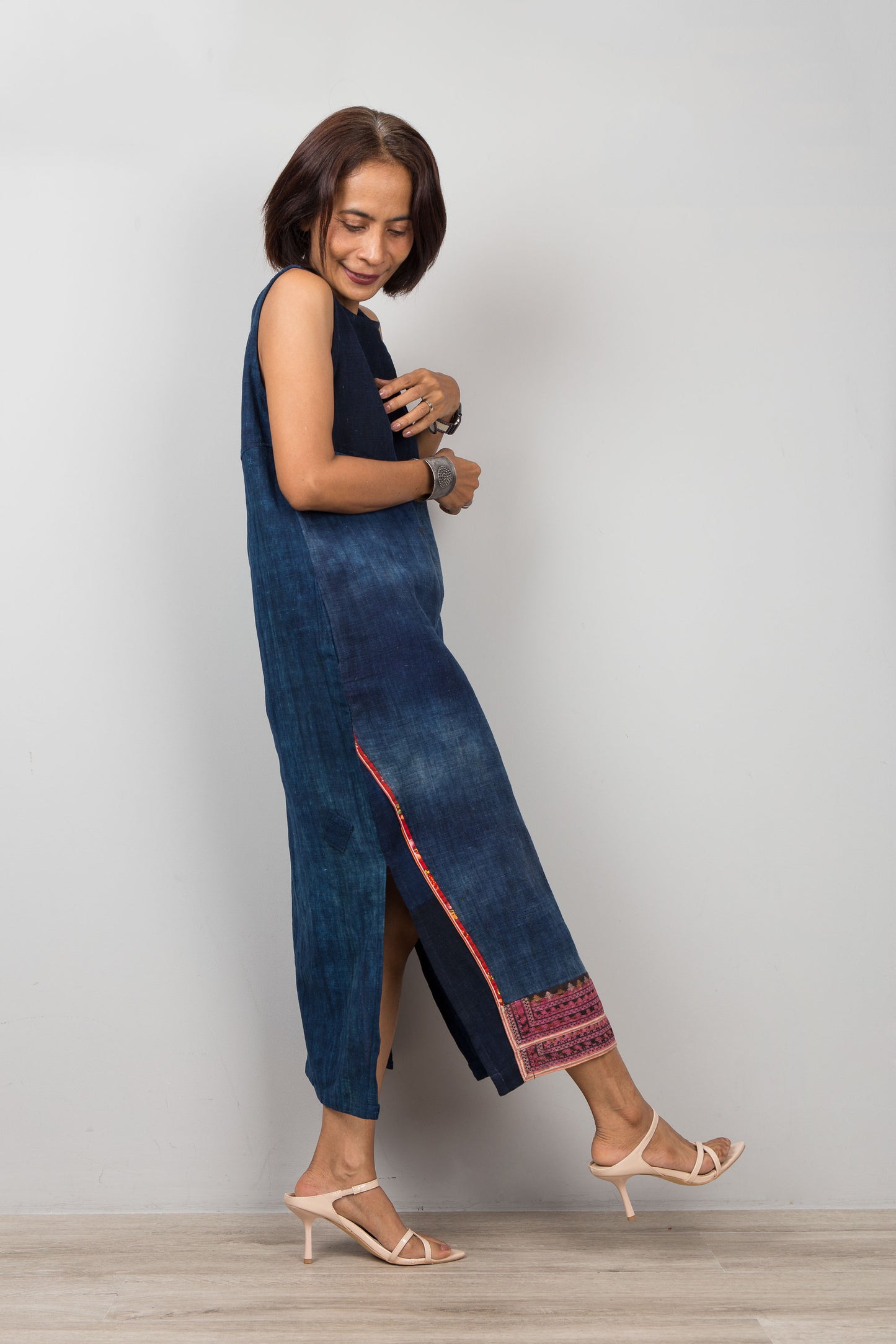 Woman wearing a blue sleeveless midi dress with embroidery, pockets and a slit on a plain background