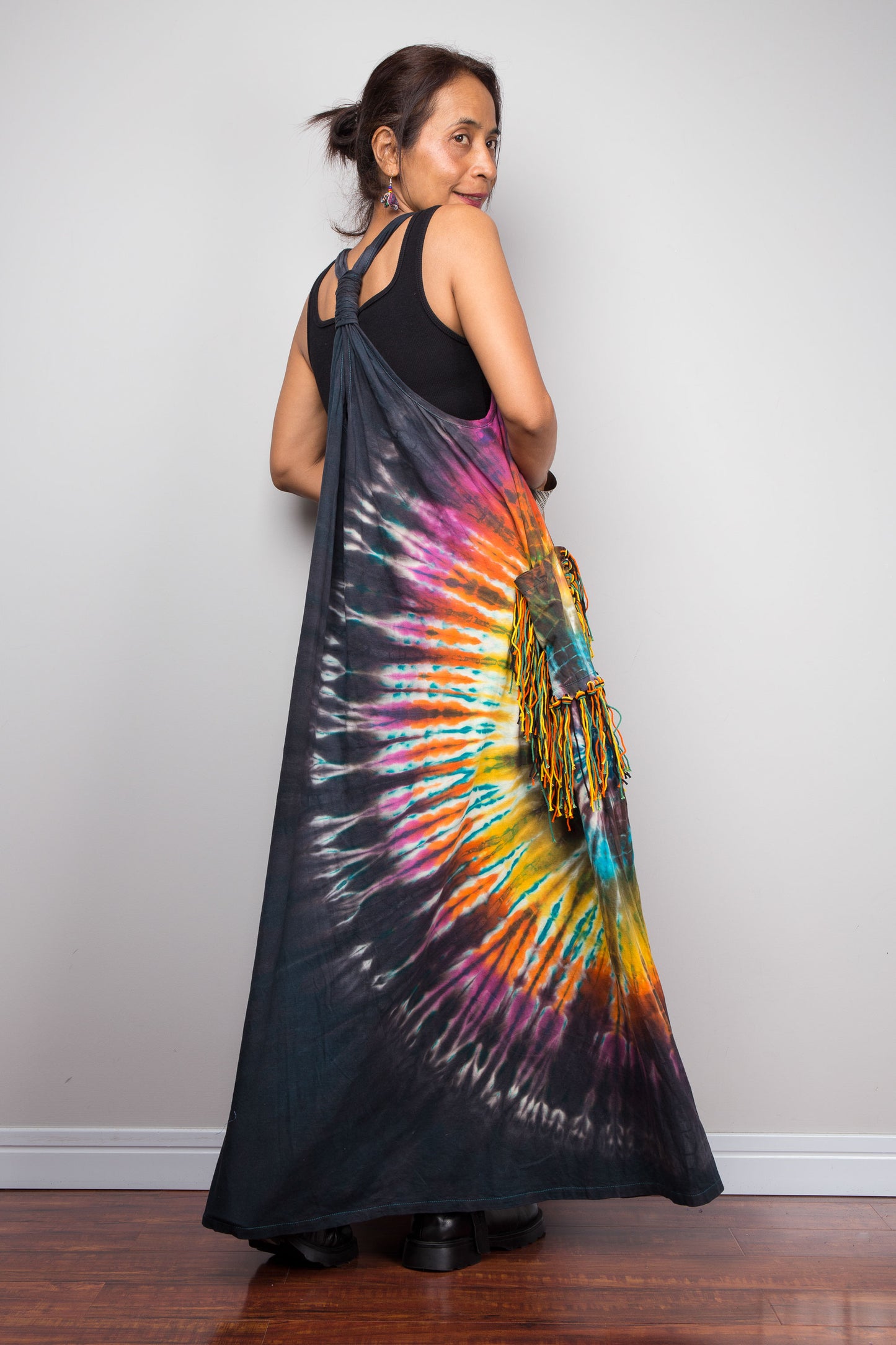 Rear side view of a model in the Nuichan tie-dye festival maxi dress, highlighting the unique twisted macramé-style gathered back strap detail, dramatic sunburst print, and fringe tassel side pockets.