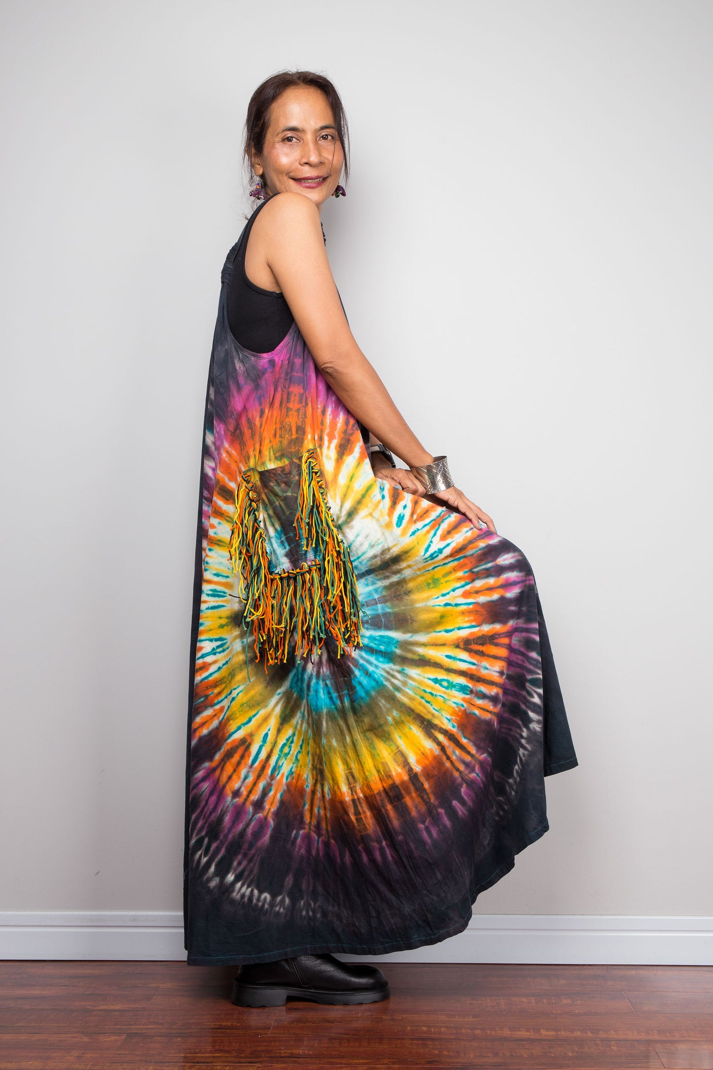 Side view of a petite model holding out the skirt of a sleeveless tie-dye maxi dress by Nuichan, showcasing the dramatic multicolour sunburst starburst pattern on a dark charcoal base with fringe tassel pockets.