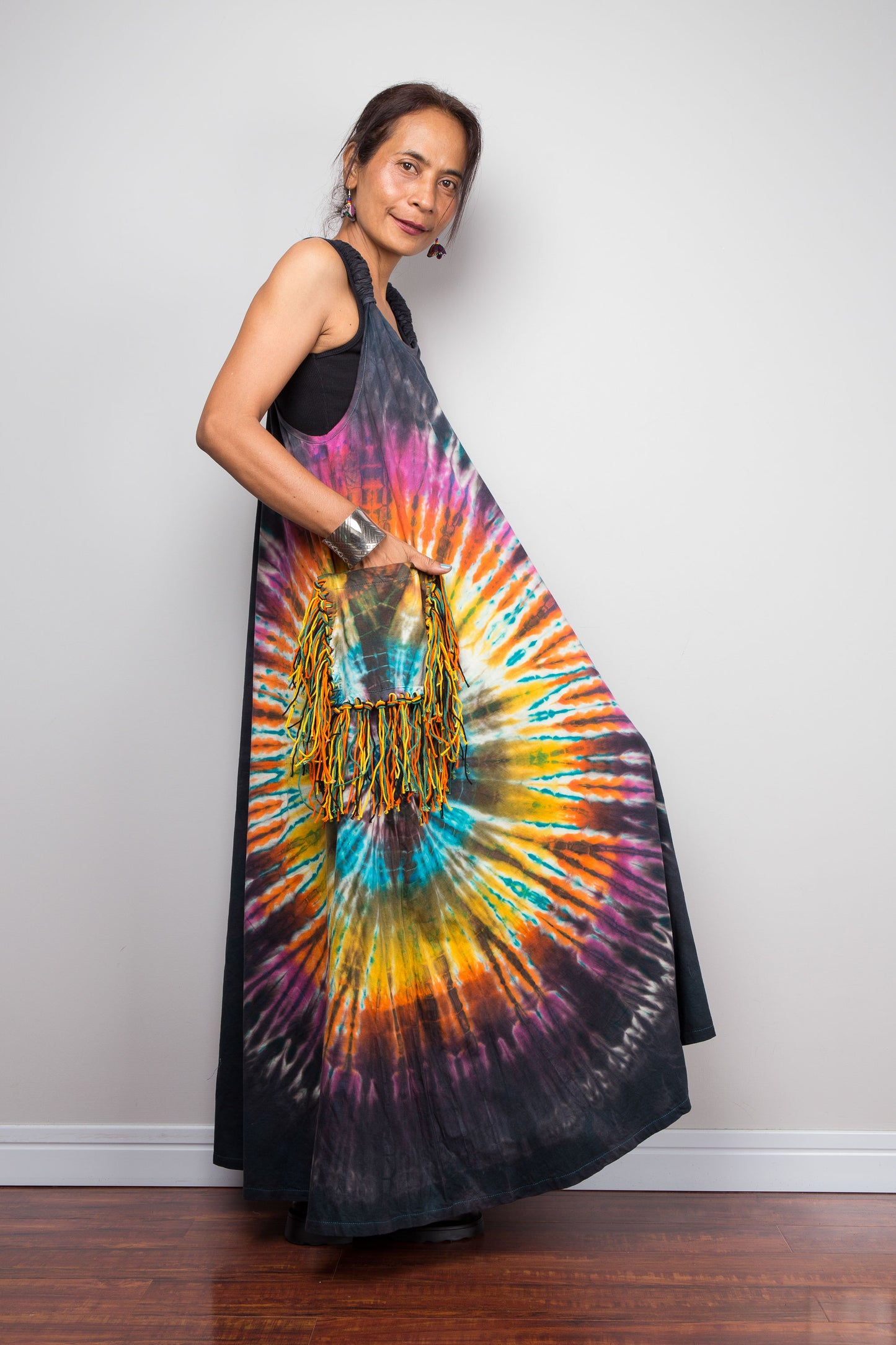 Side view of a model holding out the skirt of a sleeveless tie-dye maxi dress by Nuichan, showcasing the dramatic multicolour sunburst starburst pattern on a dark charcoal base with fringe tassel pockets.