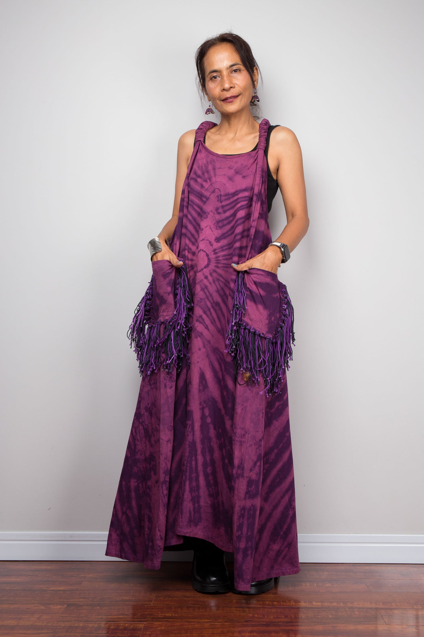Front view of a model wearing the Nuichan purple tie dye sleeveless maxi dress, hands in fringe pockets, showcasing the tonal plum and magenta starburst pattern, scoop neckline, and purple yarn tassel patch pockets.