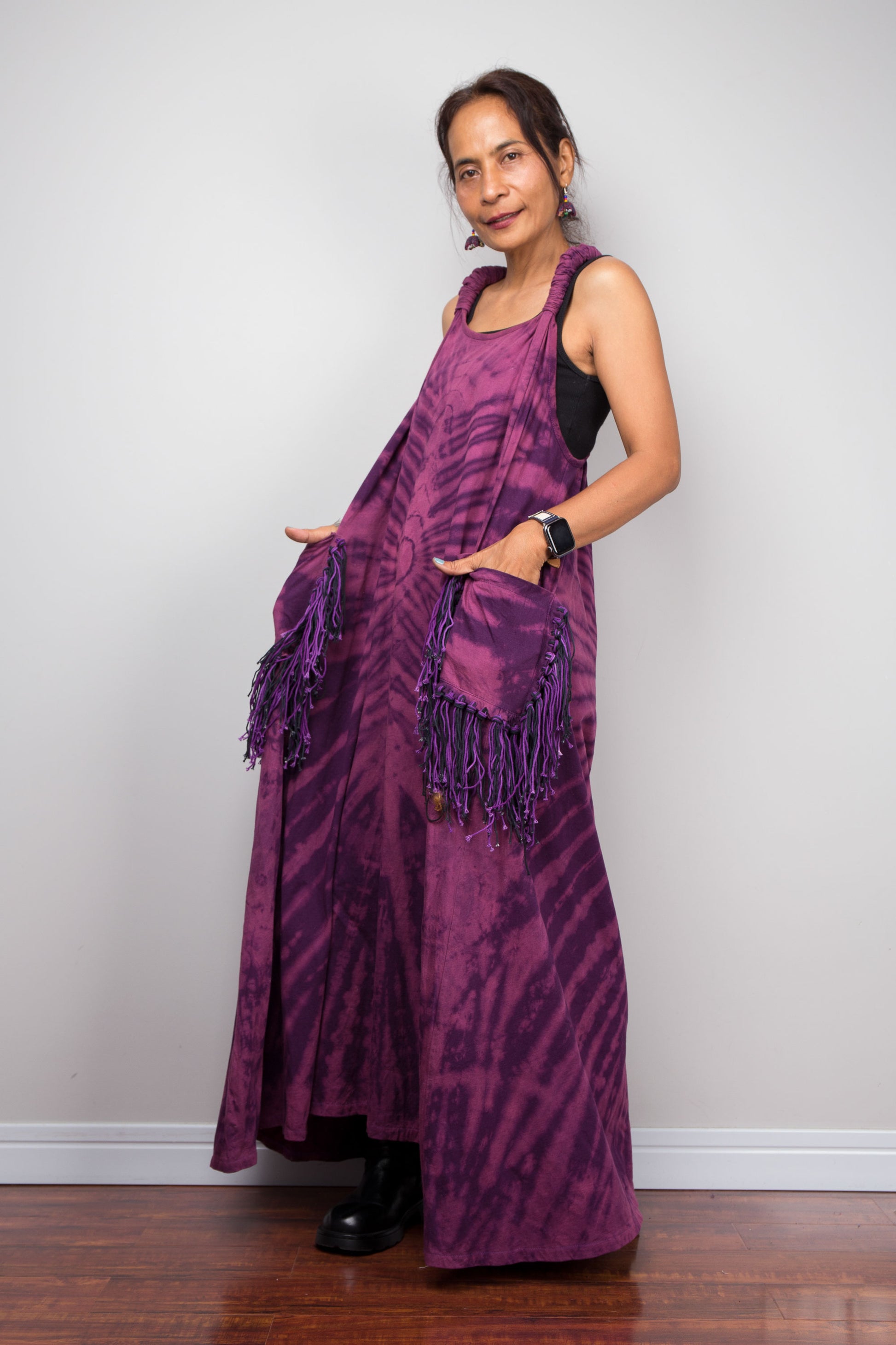 Side view of a model wearing the Nuichan purple tie dye sleeveless maxi dress, hands in fringe pockets, showcasing the tonal plum and magenta starburst pattern, scoop neckline, and purple yarn tassel patch pockets.