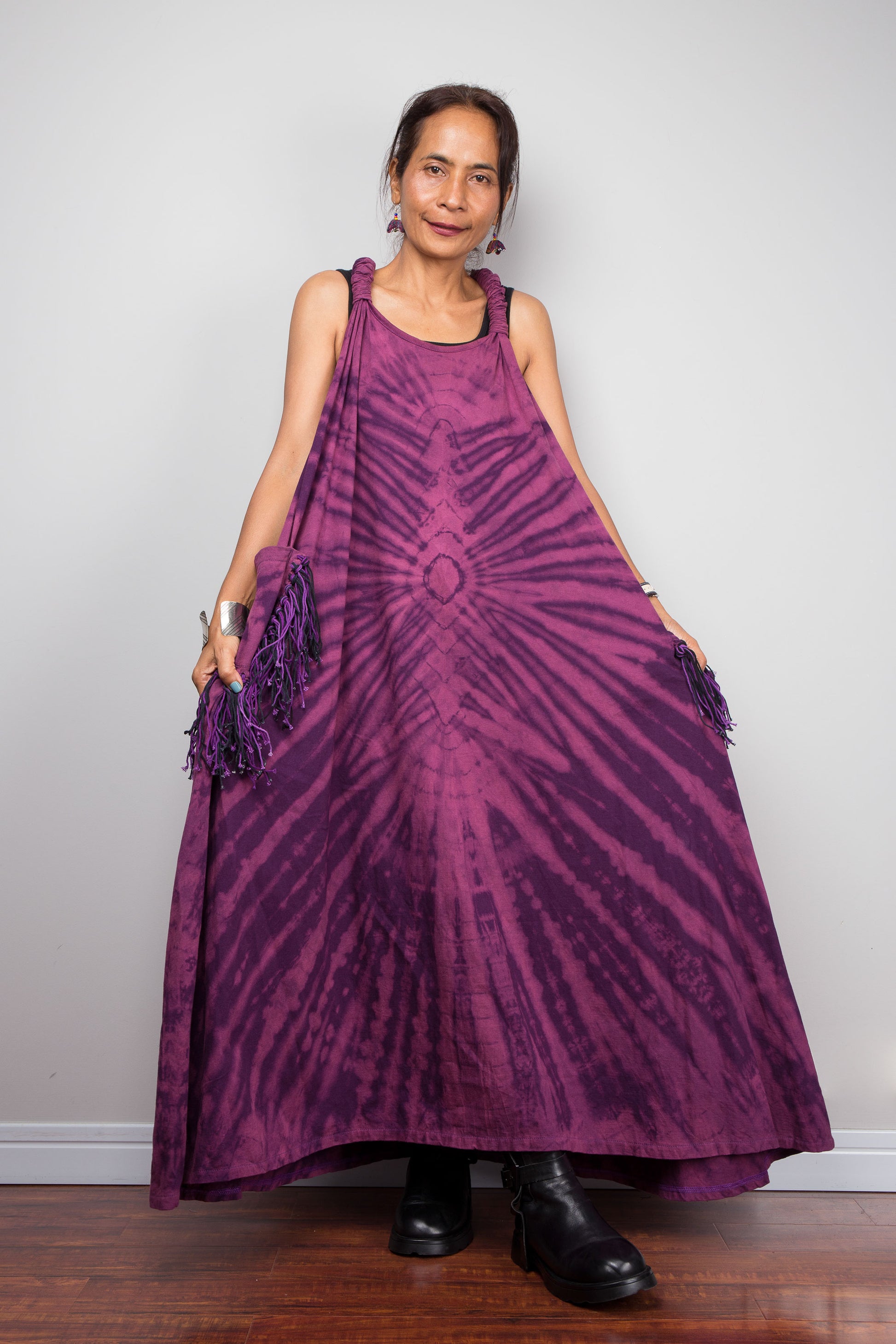 Front view of a model holding out the skirt of the Nuichan purple tie dye maxi dress, displaying the full voluminous A-line silhouette, dramatic starburst radiating from a central diamond point, and tonal purple fringe pockets.