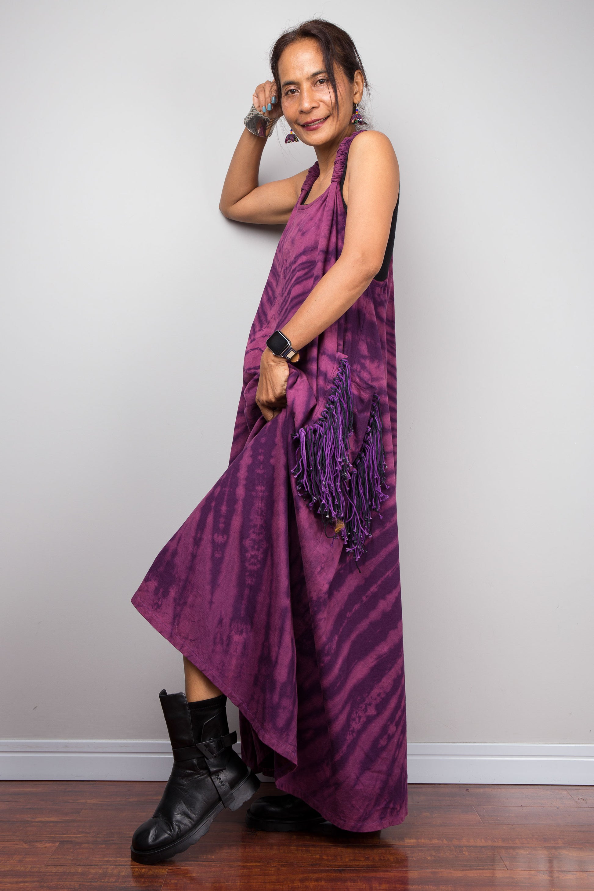 side view of a model wearing the Nuichan purple tie dye sleeveless maxi dress,  showcasing the tonal plum and magenta starburst pattern, scoop neckline, and purple yarn tassel patch pockets.