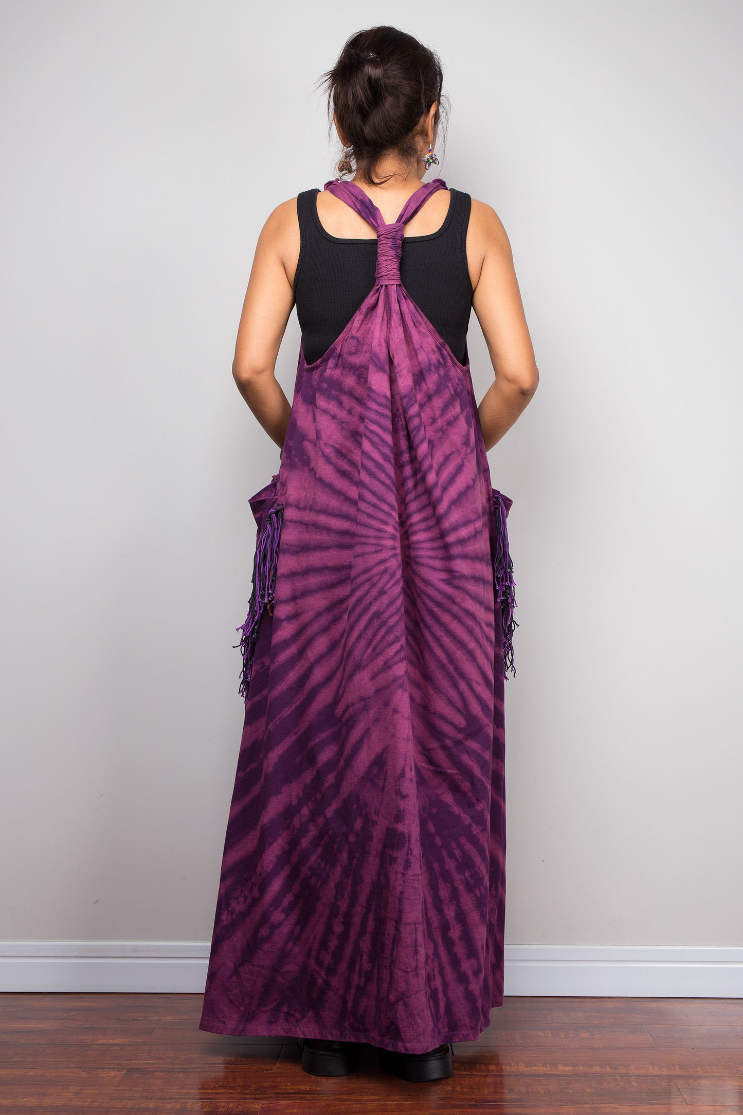 Rear view of a model in the Nuichan purple sleeveless tie dye maxi dress, highlighting the sculptural twisted macramé-style gathered back strap detail, flowing floor-length silhouette, and tonal purple fringe patch pockets.