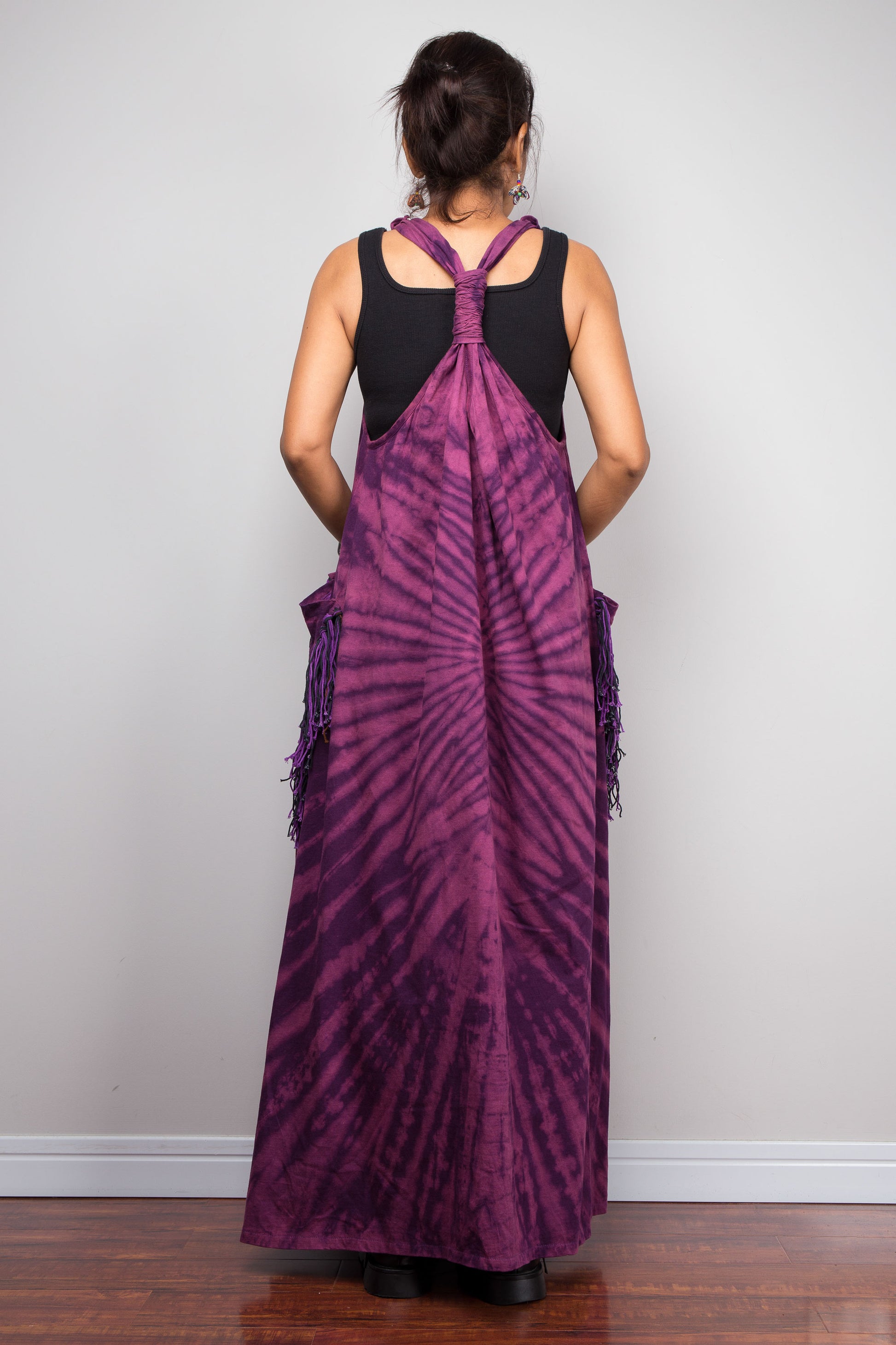 Rear view of a model in the Nuichan purple sleeveless tie dye maxi dress, highlighting the sculptural twisted macramé-style gathered back strap detail, flowing floor-length silhouette, and tonal purple fringe patch pockets.