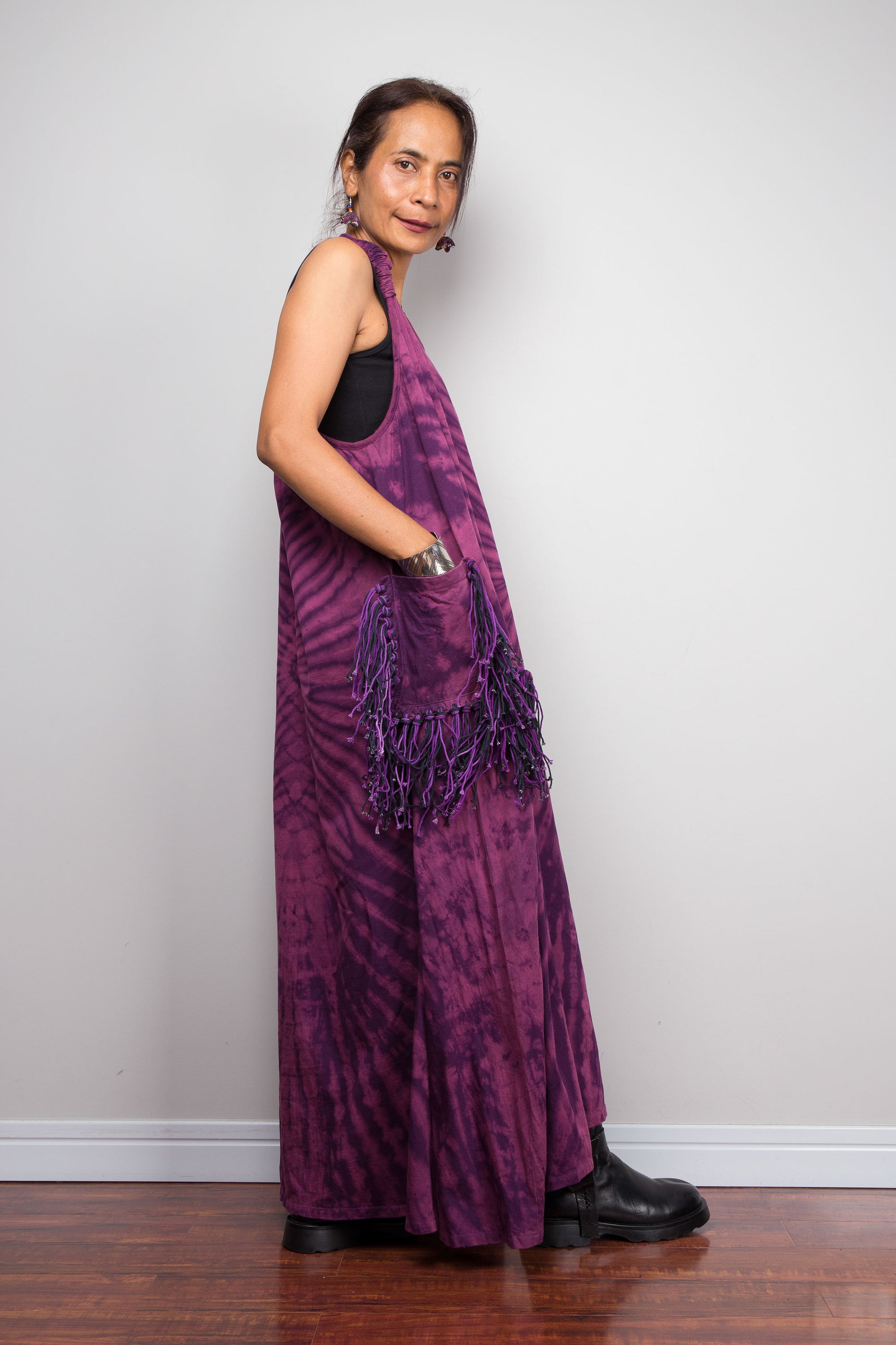 Side view of a model wearing the Nuichan purple tie dye sleeveless maxi dress, hand in pocket, showcasing the tonal plum and magenta starburst pattern and purple yarn tassel patch pockets.