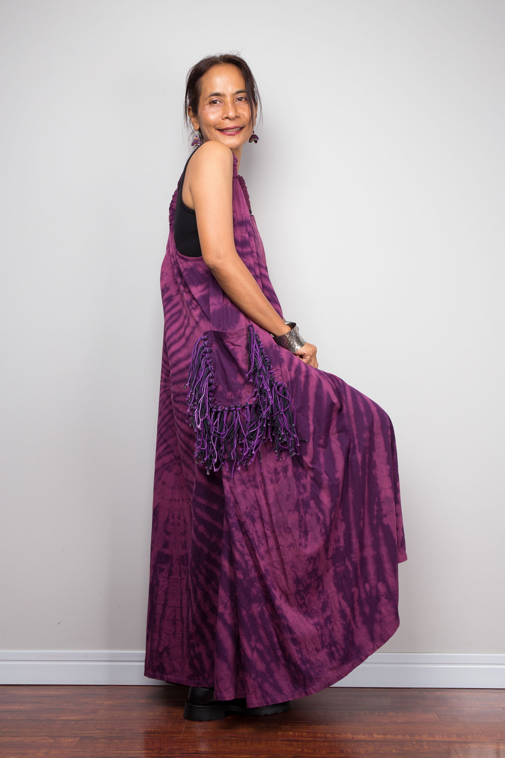 Side view of a model wearing the Nuichan purple tie dye sleeveless maxi dress, showcasing the tonal plum and magenta starburst pattern, scoop neckline, and purple yarn tassel patch pockets.