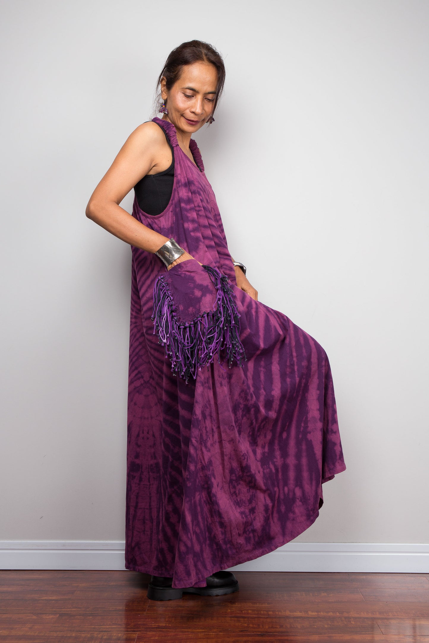 Side view of a petite model wearing the Nuichan purple tie dye sleeveless maxi dress, hand in fringe pocket, showcasing the tonal plum and magenta starburst pattern, scoop neckline, and purple yarn tassel patch pockets.