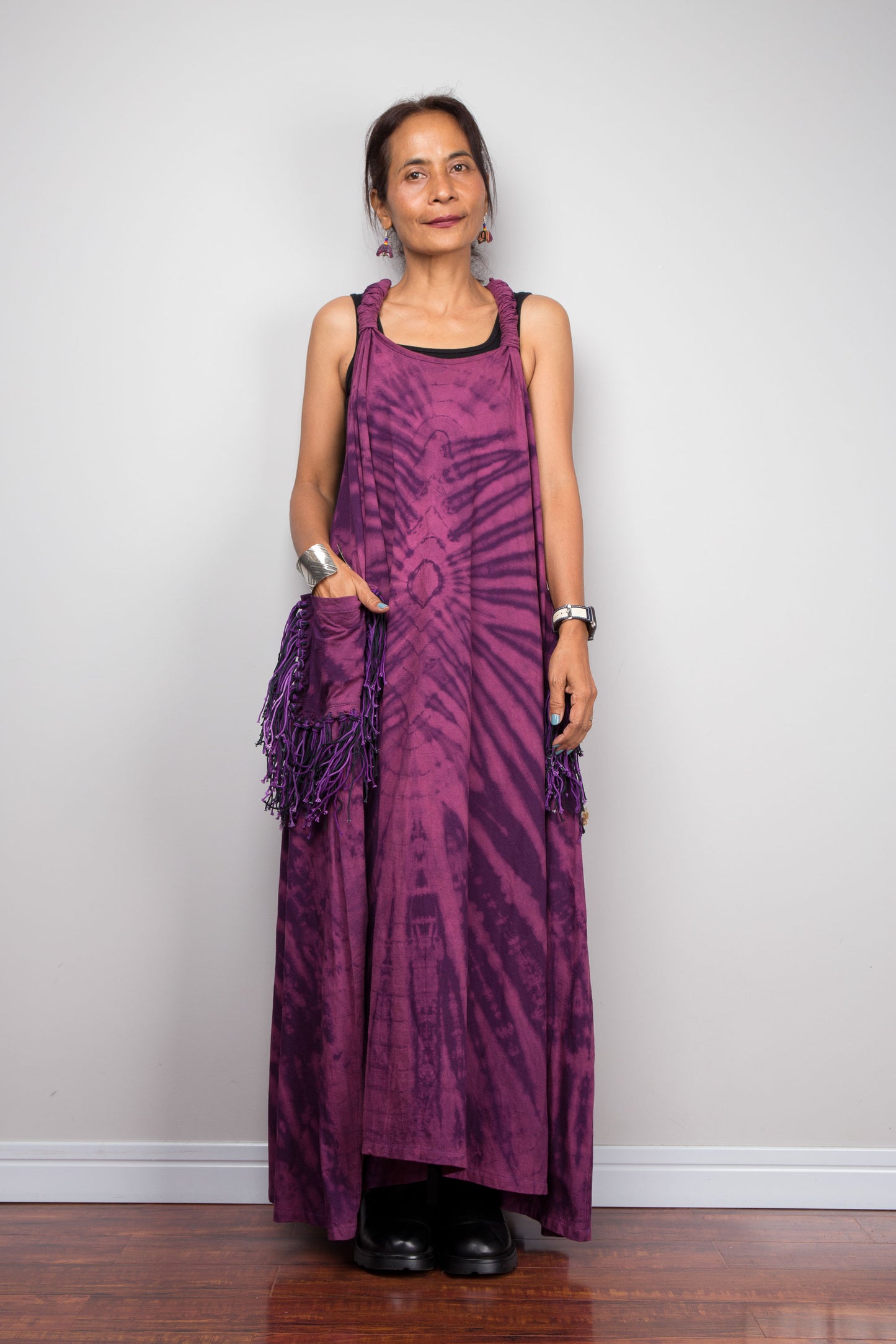 Front-facing view of a petite model wearing the Nuichan purple tie dye sleeveless maxi dress, hand in fringe pocket, showcasing the tonal plum and magenta starburst pattern, scoop neckline, and purple yarn tassel patch pockets.