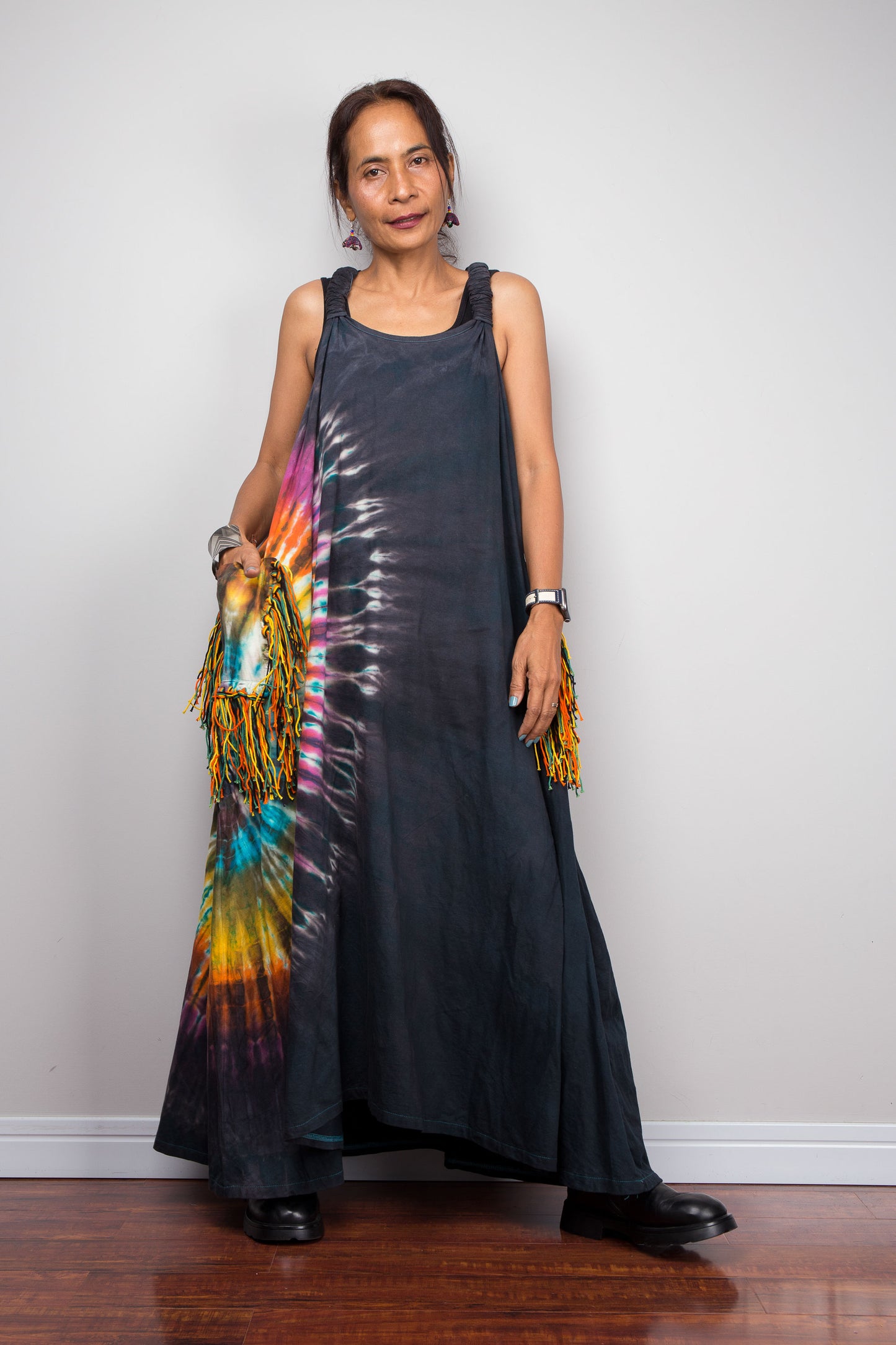 Woman wearing a long black dress with a colorful tie-dye pattern, standing against a plain white wall.