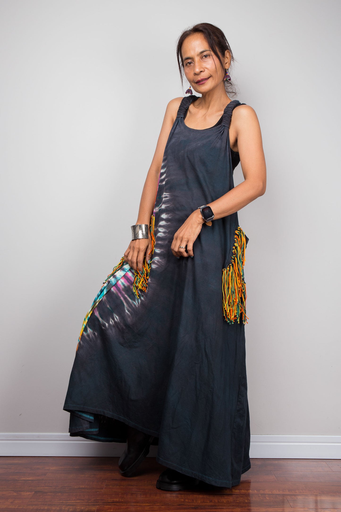 Side view of a model wearing the Nuichan sleeveless tie dye maxi dress, showing the scoop neckline, flowing A-line silhouette, and multicolour yarn fringe patch pockets on both hips.