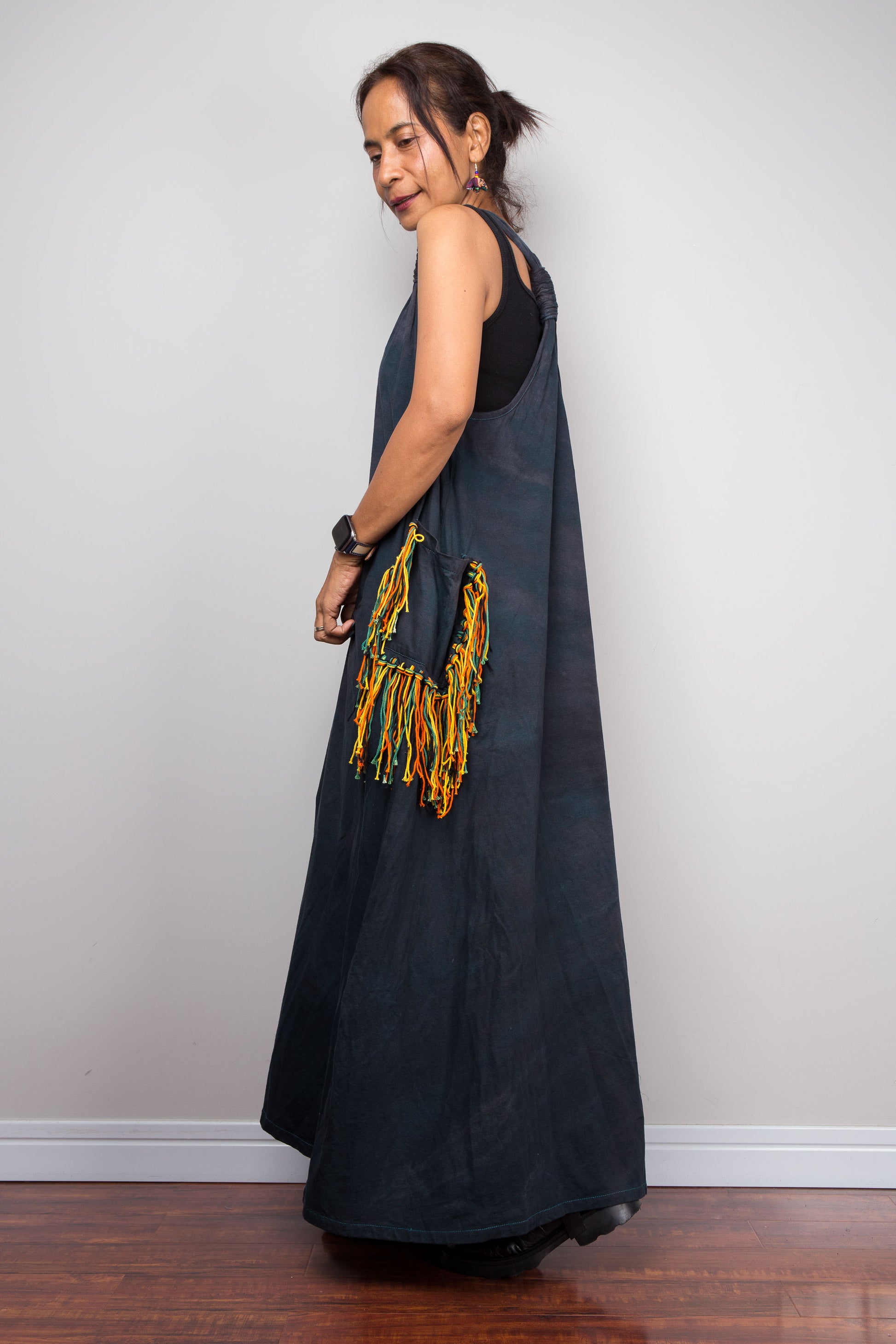 Side view of a model wearing the Nuichan sleeveless tie-dye boho maxi dress, showing the scoop neckline, flowing A-line silhouette, and a multicolour yarn fringe patch pocket on the hip.