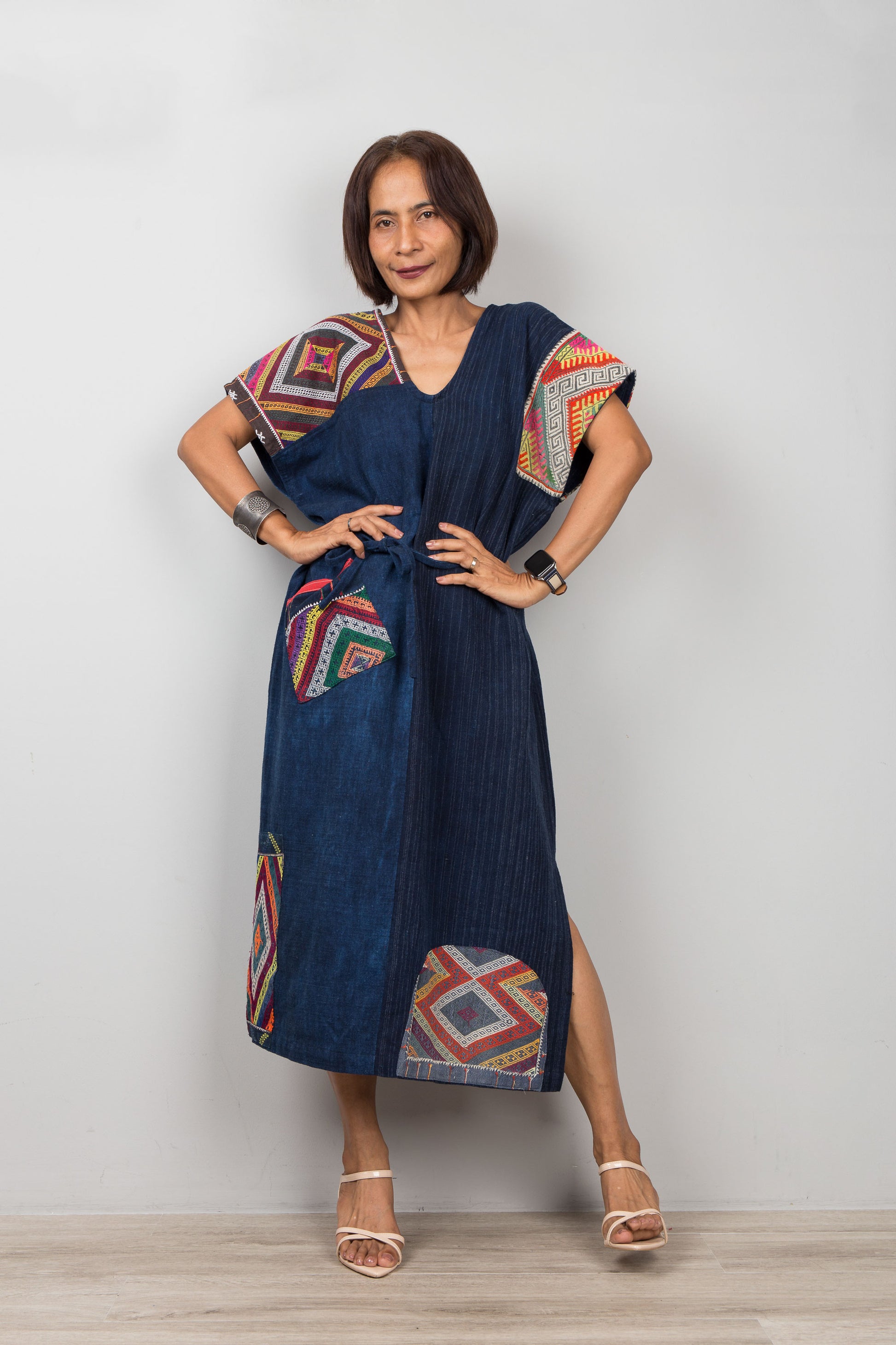 Woman wearing a blue patchwork dress with colorful geometric embroidered patterns on a plain background