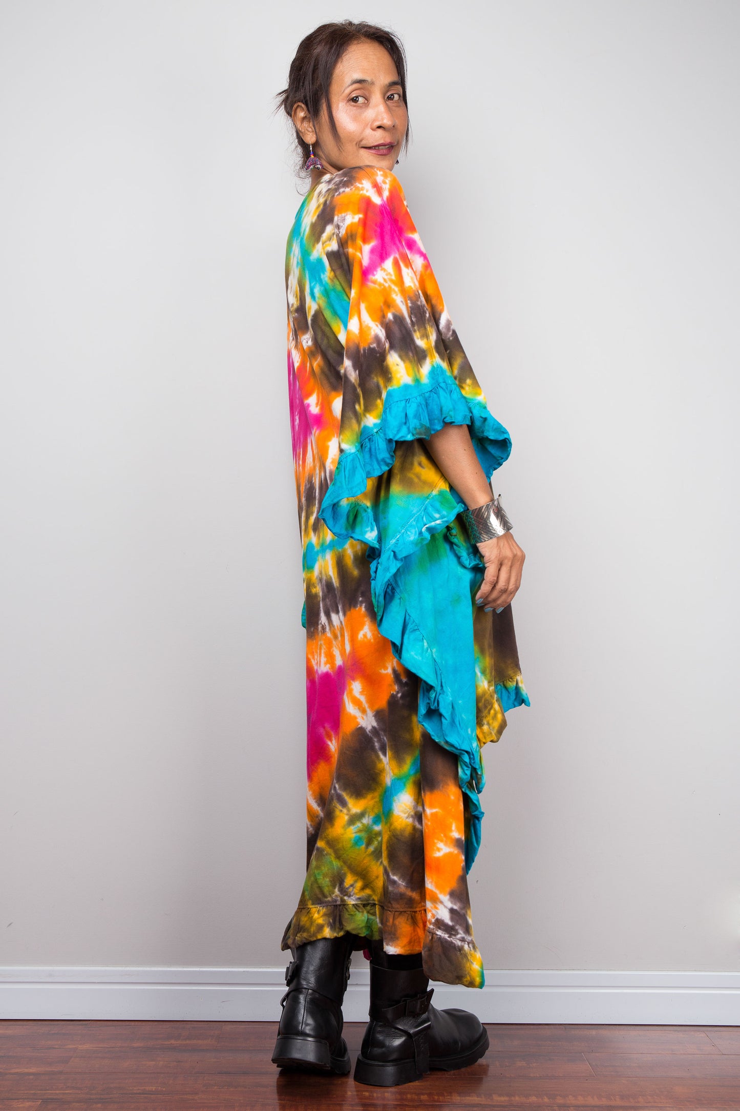 Side rear view of woman wearing a vibrant, multicolored tie-dye kaftan tunic with a high-low hemline that is short in the front and cascades into a long train at the back