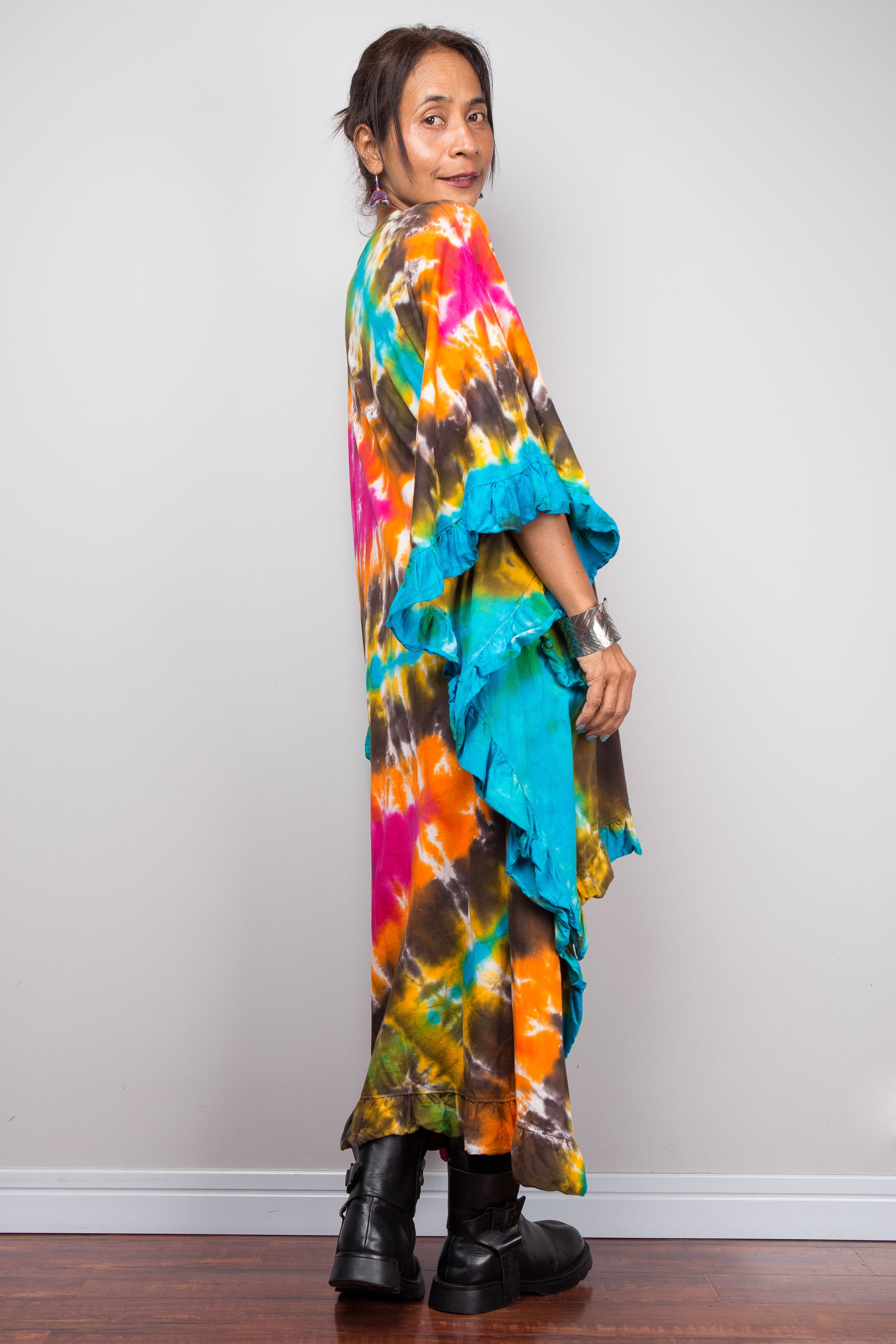 Side rear view of woman wearing a vibrant, multicolored tie-dye kaftan tunic with a high-low hemline that is short in the front and cascades into a long train at the back