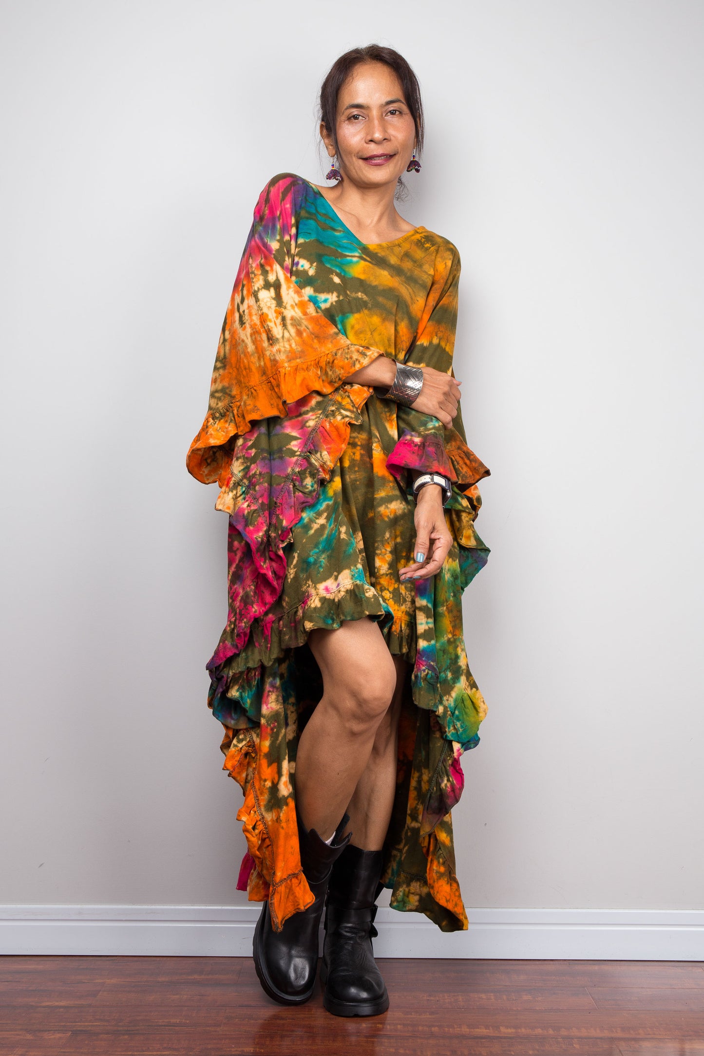 Woman wearing a colorful tie dye caftan tunic dress with black boots against a white wall.
