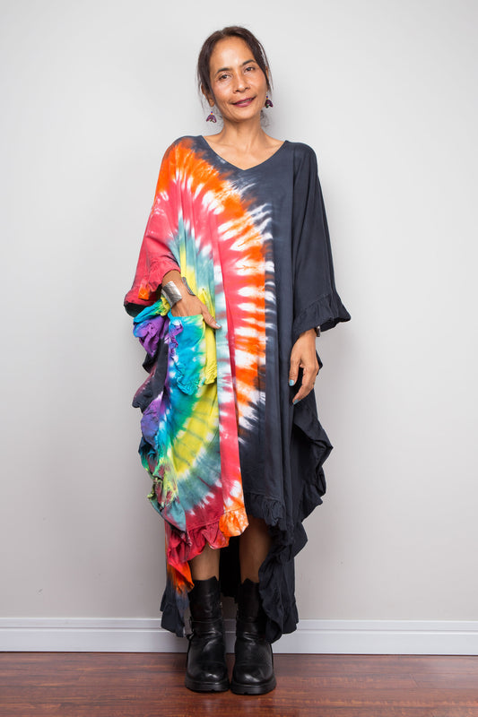 A woman modeling a long-sleeve, high-low tie-dye dress with a dark charcoal base and a vibrant multicolor spiral print, paired with black boots, standing against a plain light gray wall.