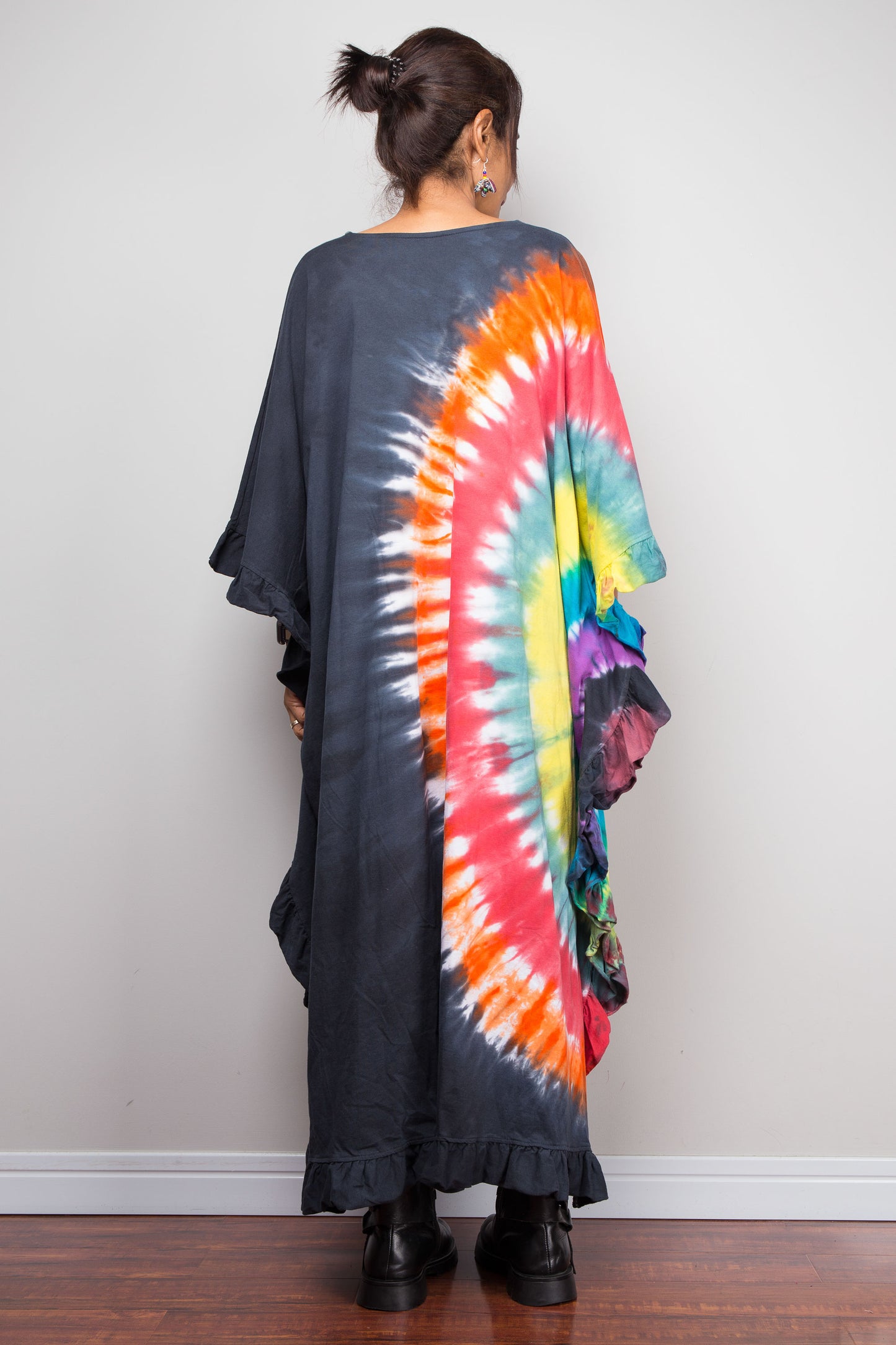 A woman viewed from behind wearing a loose, long-sleeve black dress with a large rainbow tie-dye spiral pattern on the back, standing on a wooden floor against a plain light gray wall.