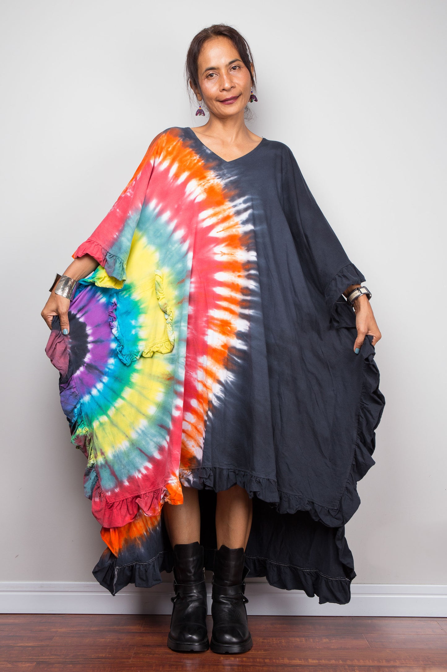 A woman modeling a black ruffled high-low dress with a vibrant rainbow tie-dye pattern draped over one shoulder, standing against a plain light gray wall.