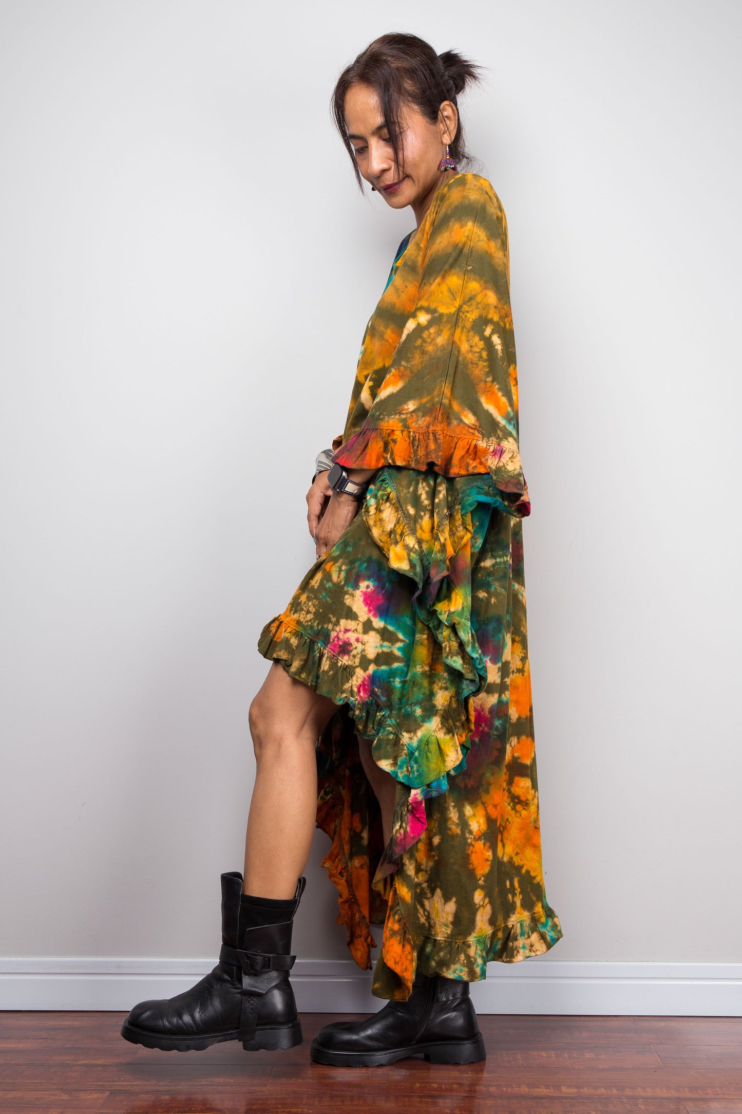 Person wearing a colorful high-low tie dye caftan tunic dress with black boots against a white wall.