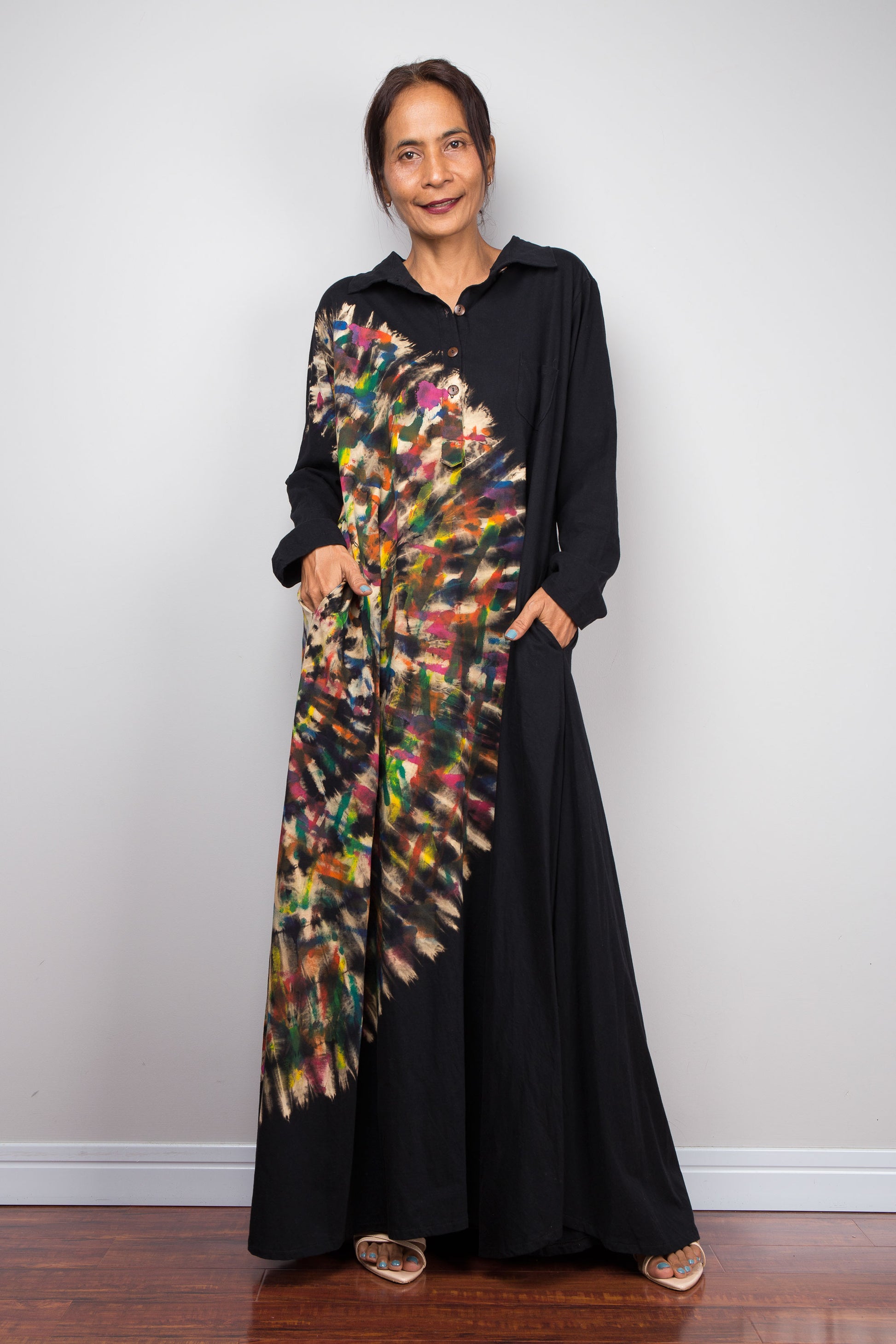 Front of woman in black long-sleeve maxi shirt dress. An asymmetric, vibrant hand-dyed multi-color tie-dye sunburst sweeps diagonally across the front. She has hands in side pockets.