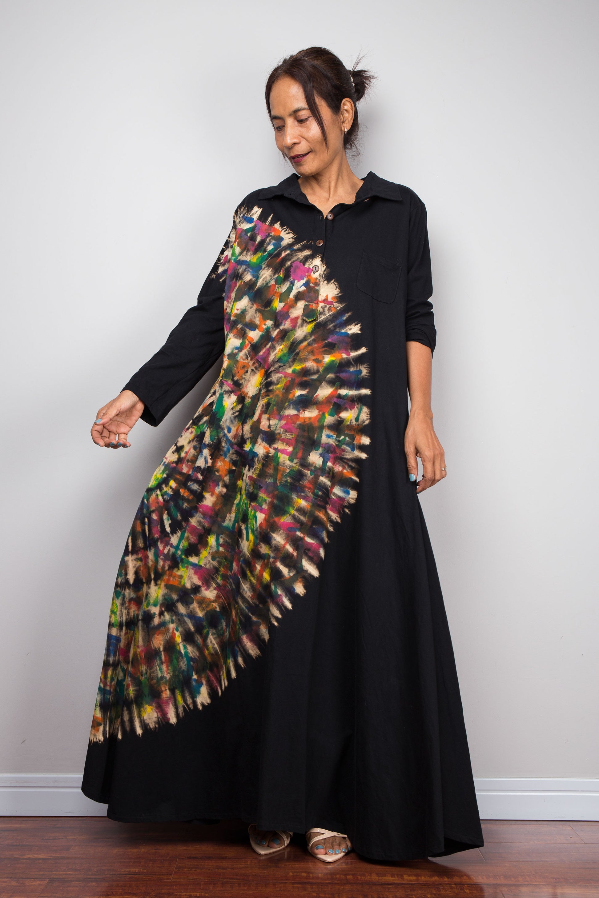 Petite woman in black long-sleeve tie dye maxi shirt dress. An asymmetric, vibrant hand-dyed multi-color tie-dye sunburst sweeps diagonally across the front. She has hands in side pockets.