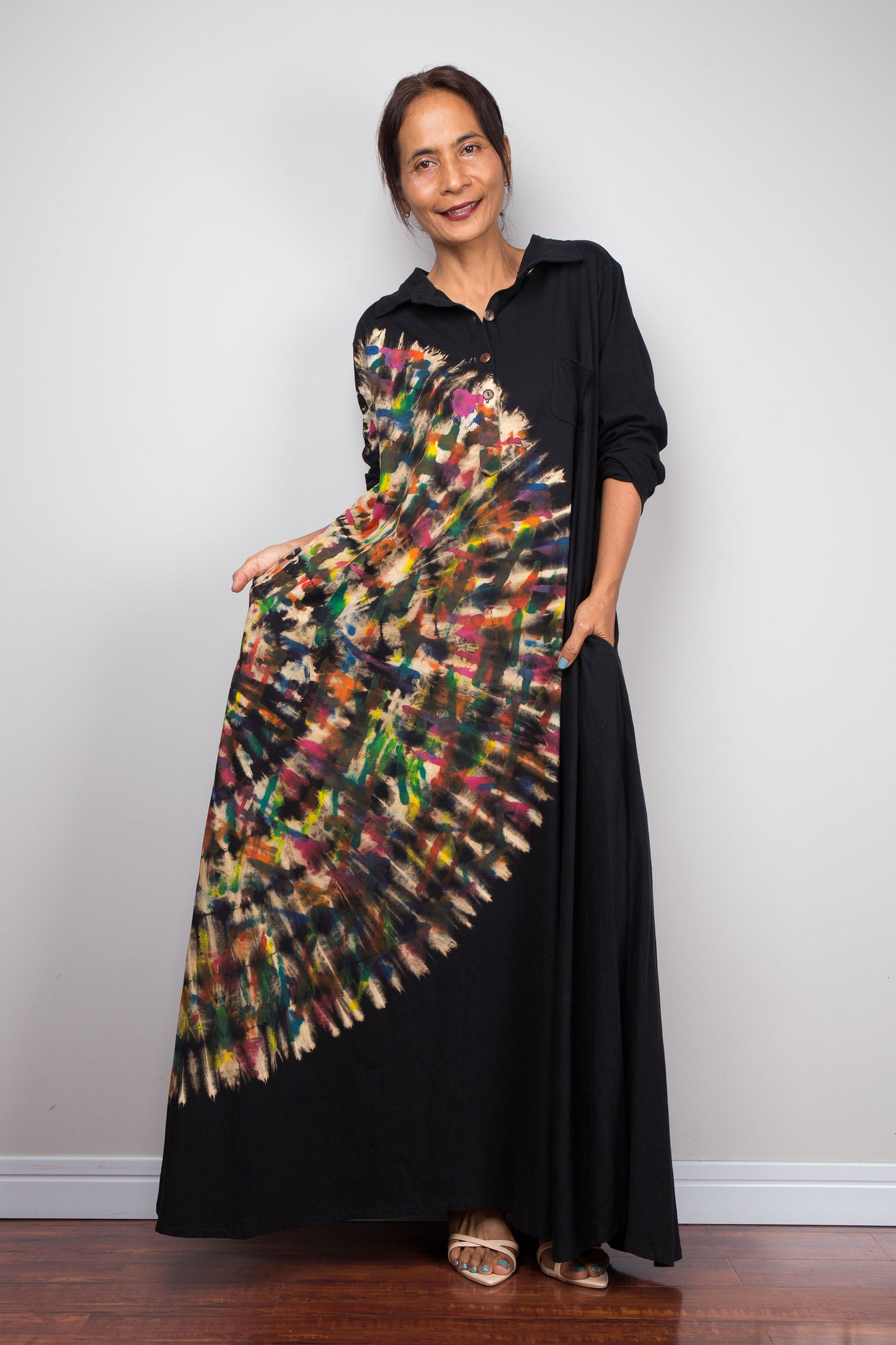 Front view of woman in black long-sleeve tie dye maxi shirt dress. An asymmetric, vibrant hand-dyed multi-color tie-dye sunburst sweeps diagonally across the front. She has hands in side pockets.
