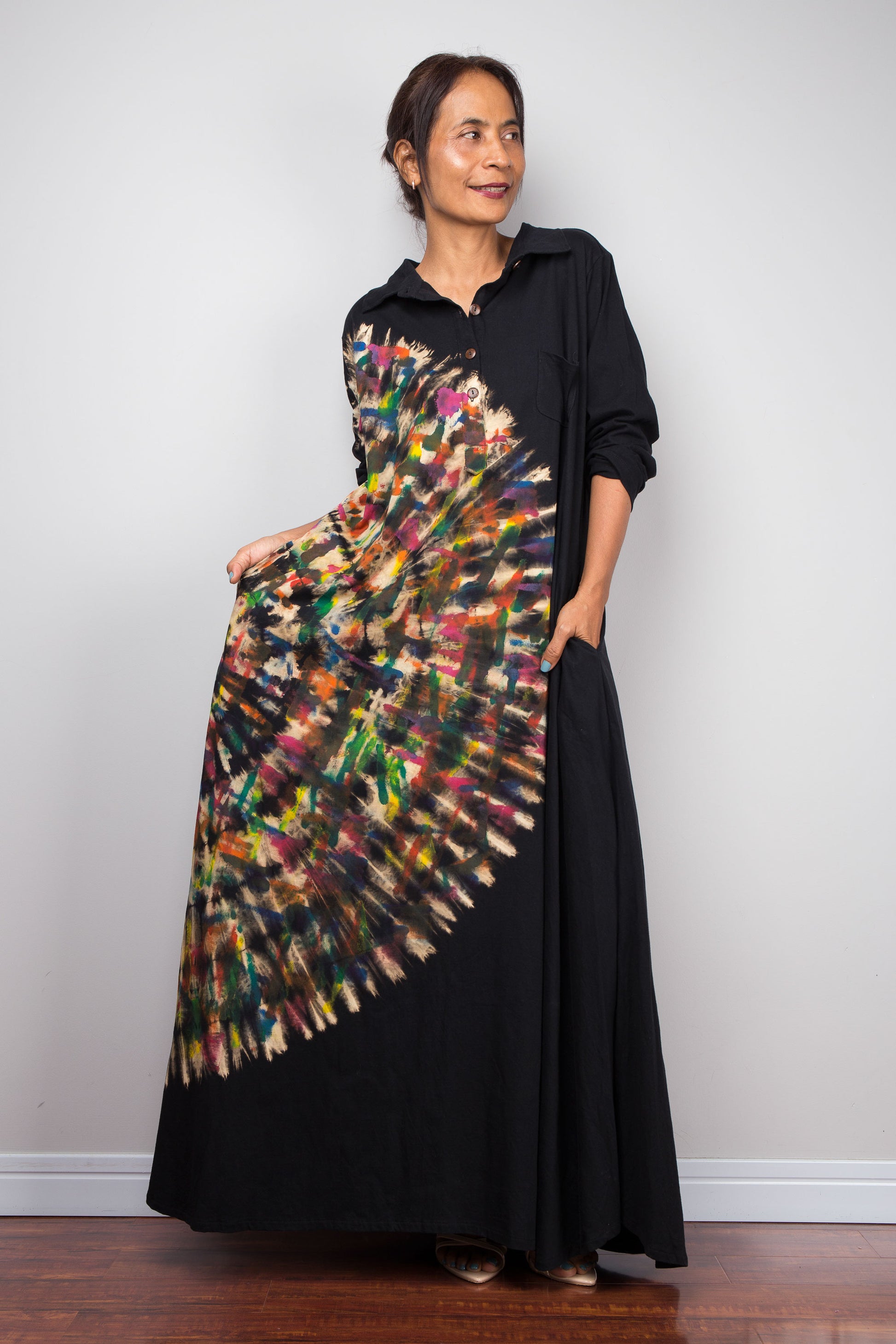 Front view of petite woman in a black long-sleeve tie dye maxi shirt dress. An asymmetric, vibrant hand-dyed multi-color tie-dye sunburst sweeps diagonally across the front. She has hands in side pockets.