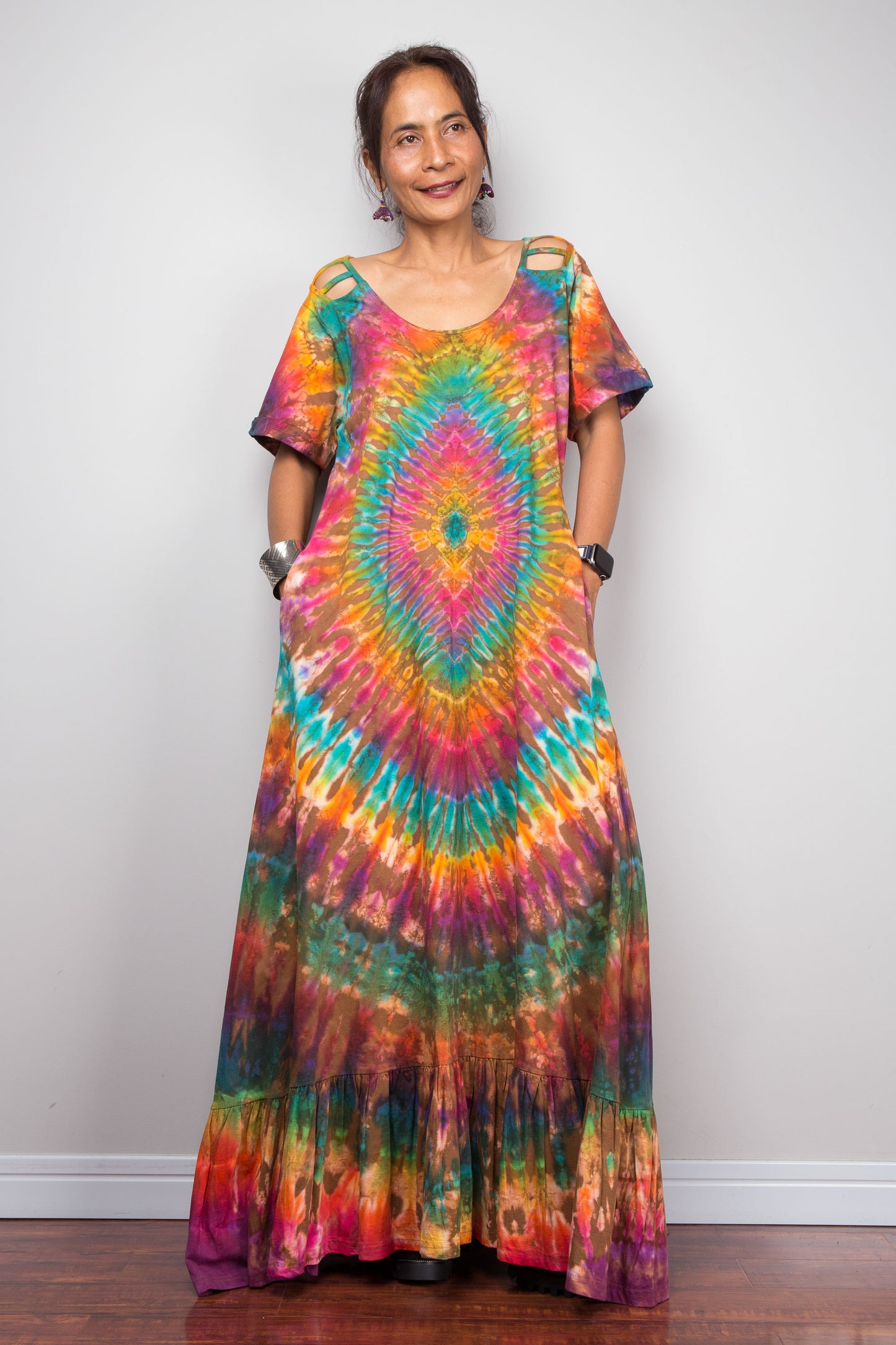 Front-facing view of a model wearing the Nuichan rainbow tie dye maxi dress with hands in side pockets, showcasing the full-spectrum starburst pattern radiating from a central teal diamond, scoop neckline, short sleeves with cut-out ladder slash details, and tiered ruffle hem.