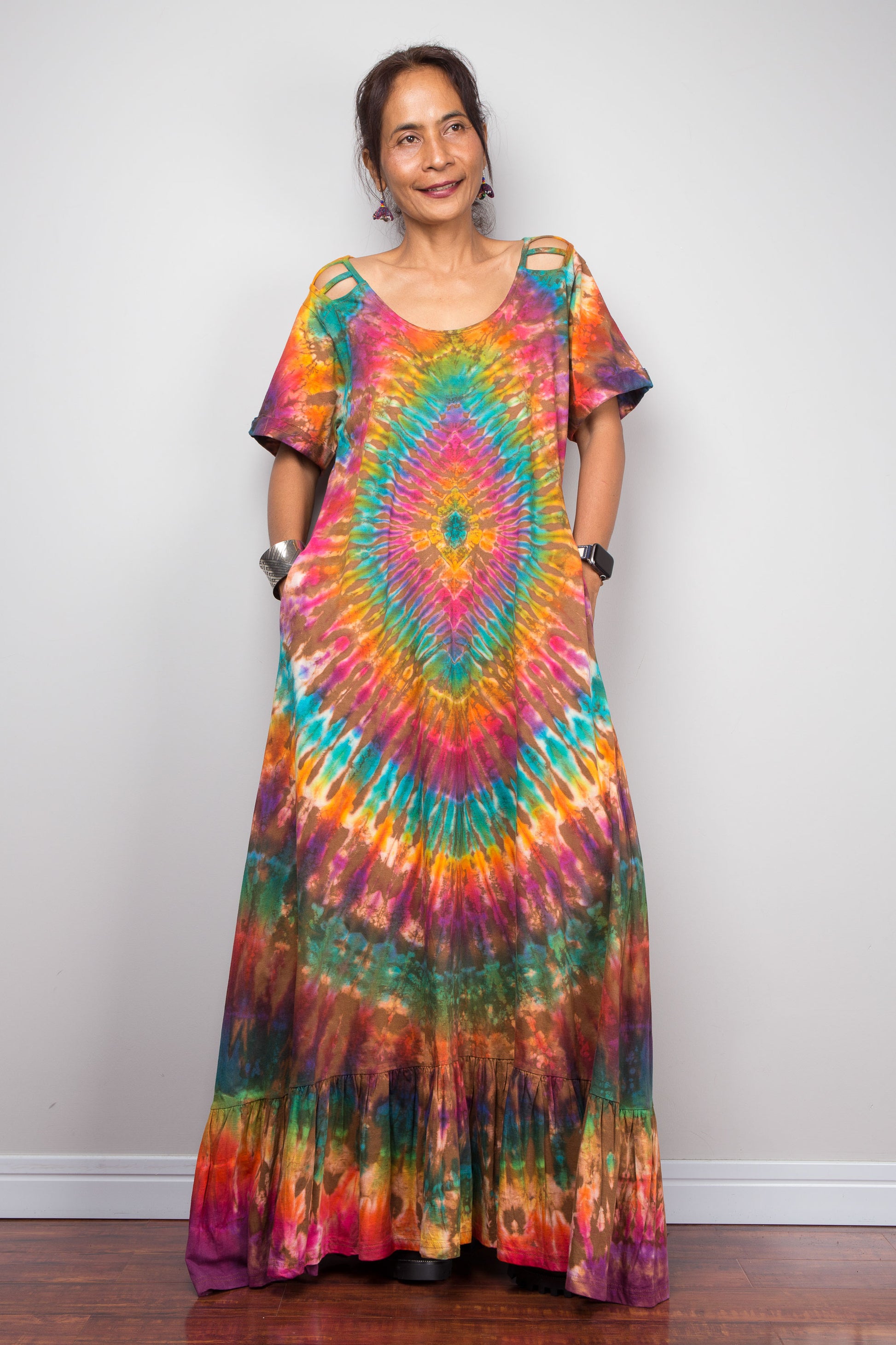 Front-facing view of a model wearing the Nuichan rainbow tie dye maxi dress with hands in side pockets, showcasing the full-spectrum starburst pattern radiating from a central teal diamond, scoop neckline, short sleeves with cut-out ladder slash details, and tiered ruffle hem.