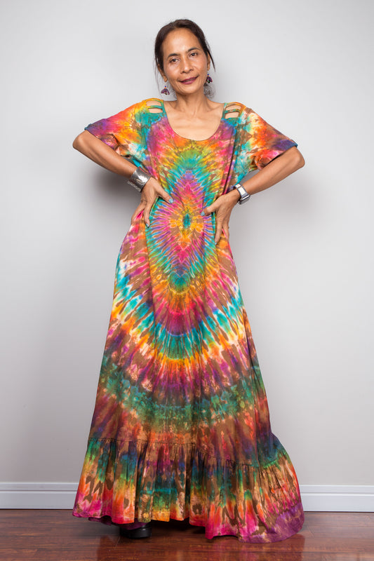 Front-facing view of a model wearing the Nuichan rainbow tie dye maxi dress with hands on the hips, showcasing the full-spectrum starburst pattern radiating from a central teal diamond, scoop neckline, short sleeves with cut-out ladder slash details, and tiered ruffle hem.