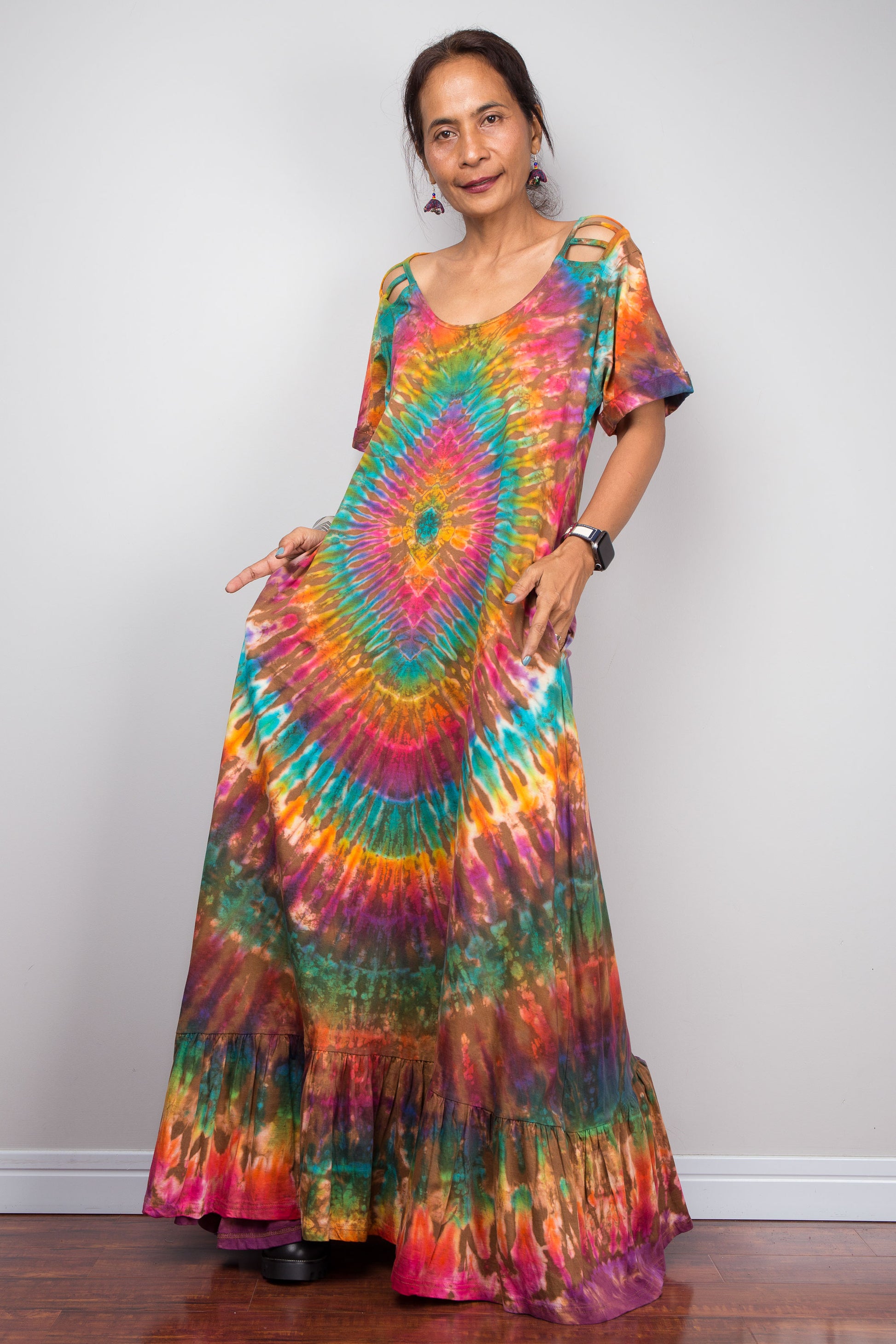 Three-quarter side view of a model wearing the Nuichan rainbow tie dye maxi dress with hands in pockets, displaying the full A-line silhouette, kaleidoscopic full-spectrum starburst, cut-out shoulder details, and dramatic floor-sweeping tiered ruffle hem.