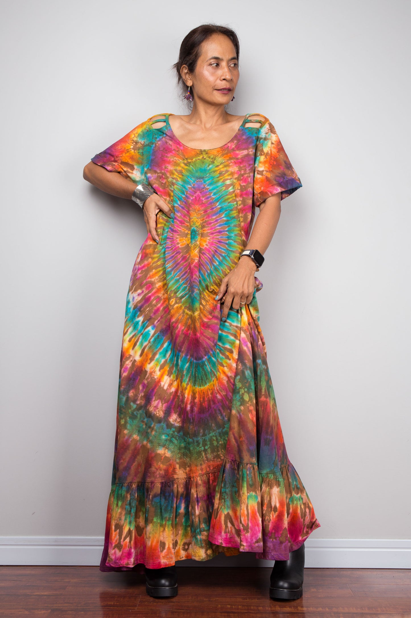 Model wearing the Nuichan rainbow tie dye maxi dress with hand on hip, showcasing the full-spectrum starburst pattern radiating from a central teal diamond, scoop neckline, short sleeves with cut-out ladder slash details, and tiered ruffle hem.