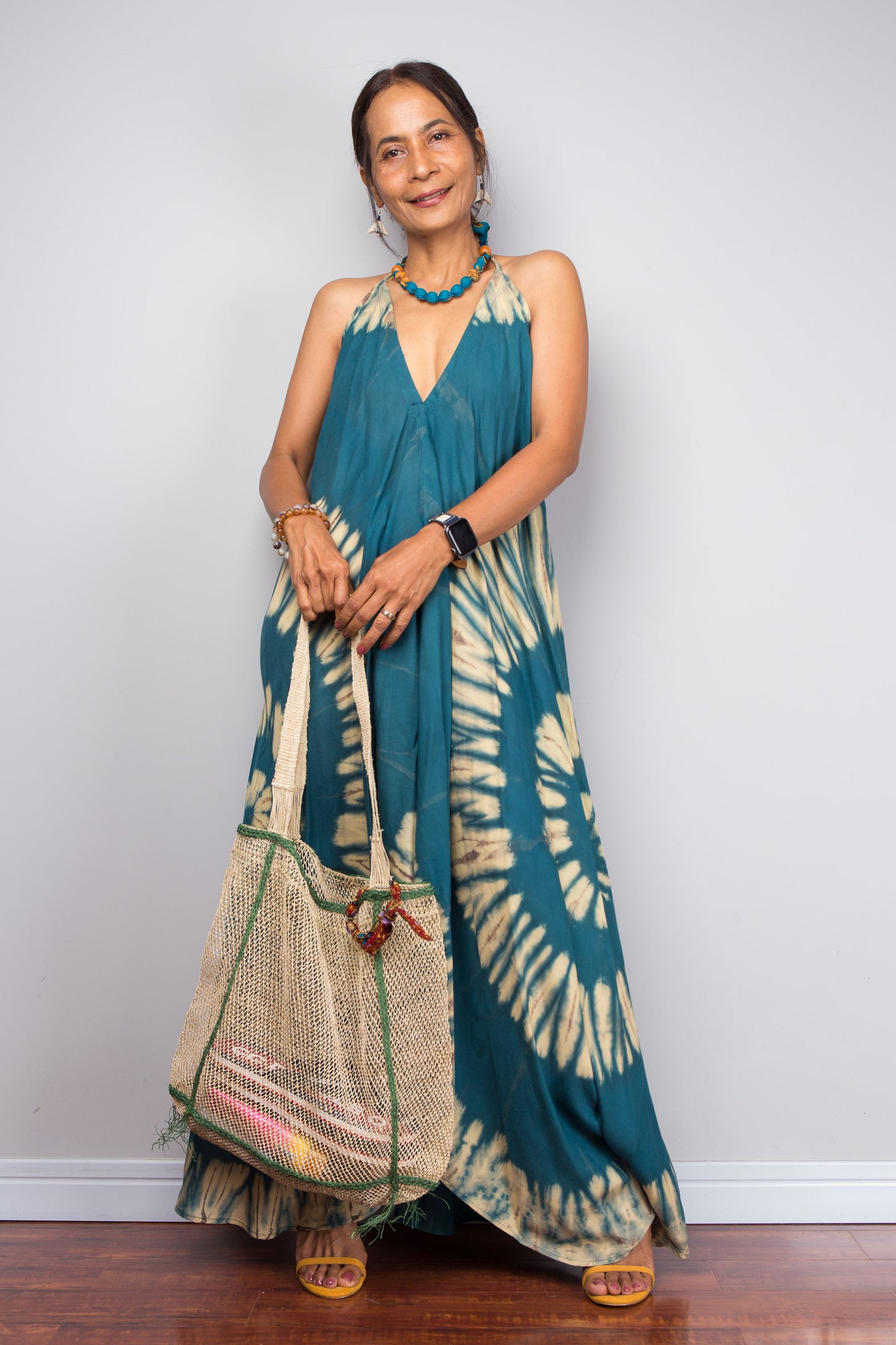 Woman wearing a long teal and beige tie-dye dress with a matching bag against a white wall.