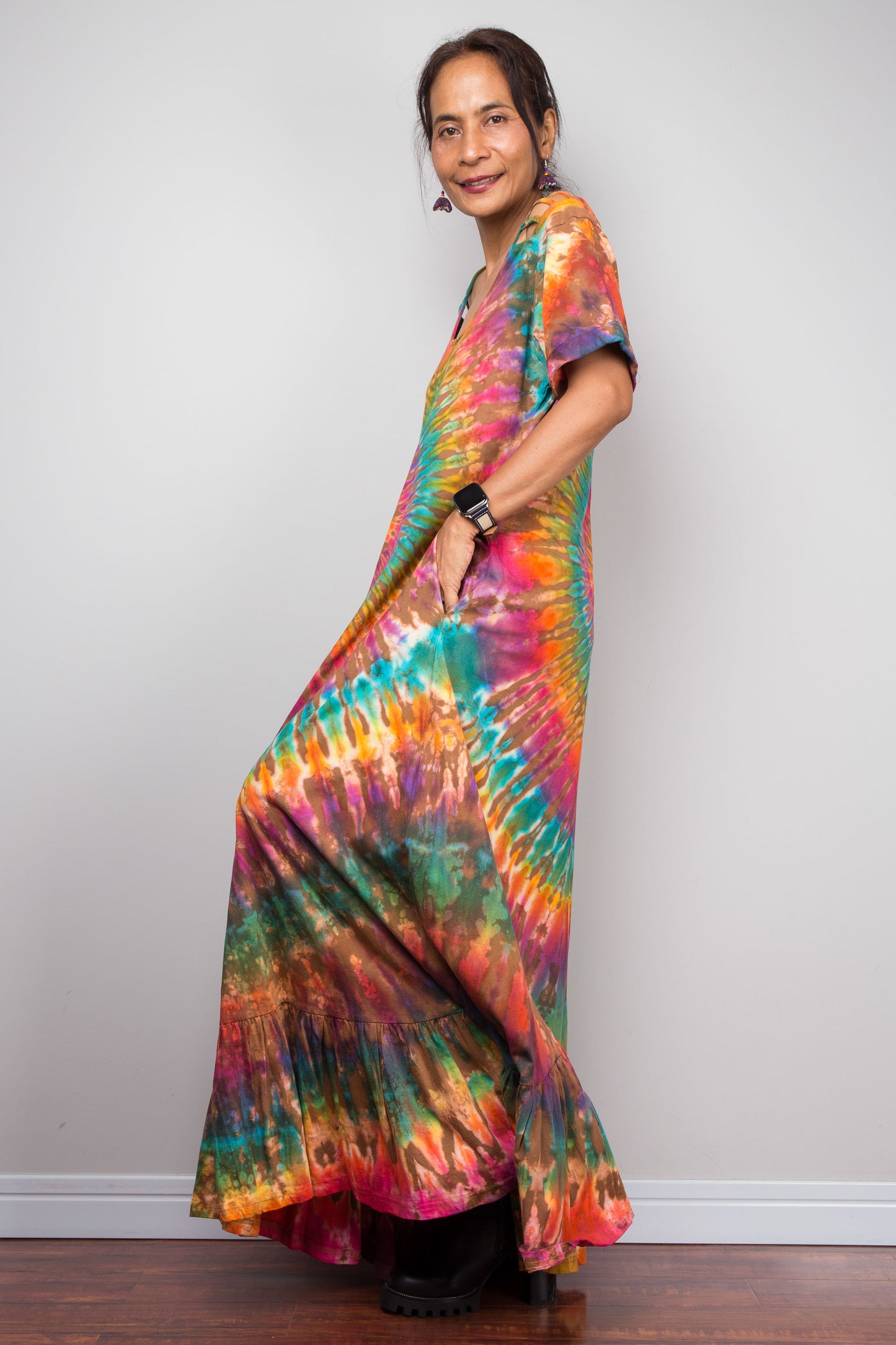 Side view of a model wearing the Nuichan rainbow tie dye maxi dress with hand in side pocket, showcasing the full-spectrum starburst pattern radiating from a central teal diamond, scoop neckline, short sleeves with cut-out ladder slash details, and tiered ruffle hem.