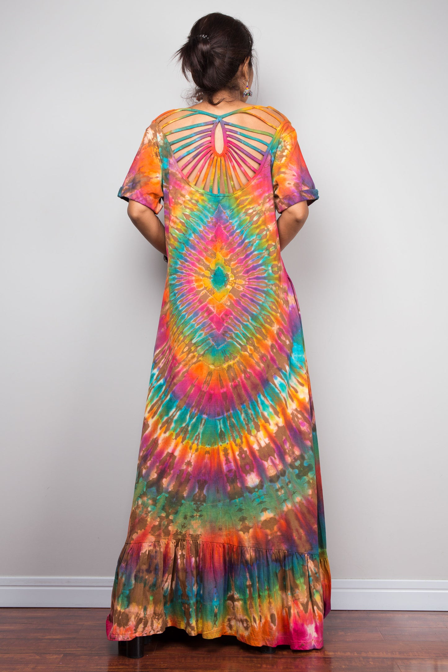 Rear view of a model in the Nuichan rainbow tie dye maxi dress, highlighting the dramatic cage-style cut-out sunburst back with rainbow-dyed fabric strips radiating from a central keyhole, full-spectrum starburst pattern, and tiered ruffle hem.