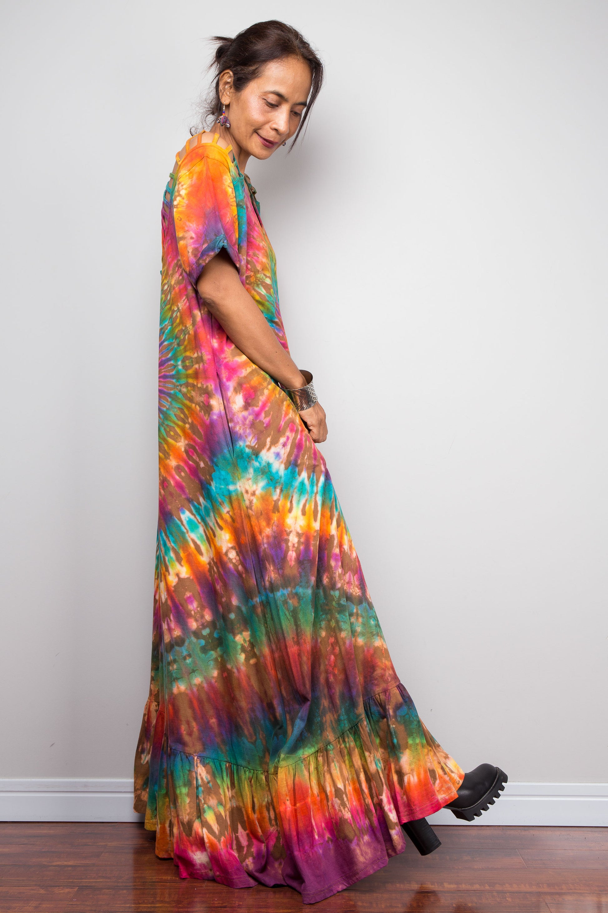 Side view of a model wearing the Nuichan rainbow tie dye maxi dress, showcasing the full-spectrum starburst pattern radiating from a central teal diamond, scoop neckline, short sleeves with cut-out ladder slash details, and tiered ruffle hem.
