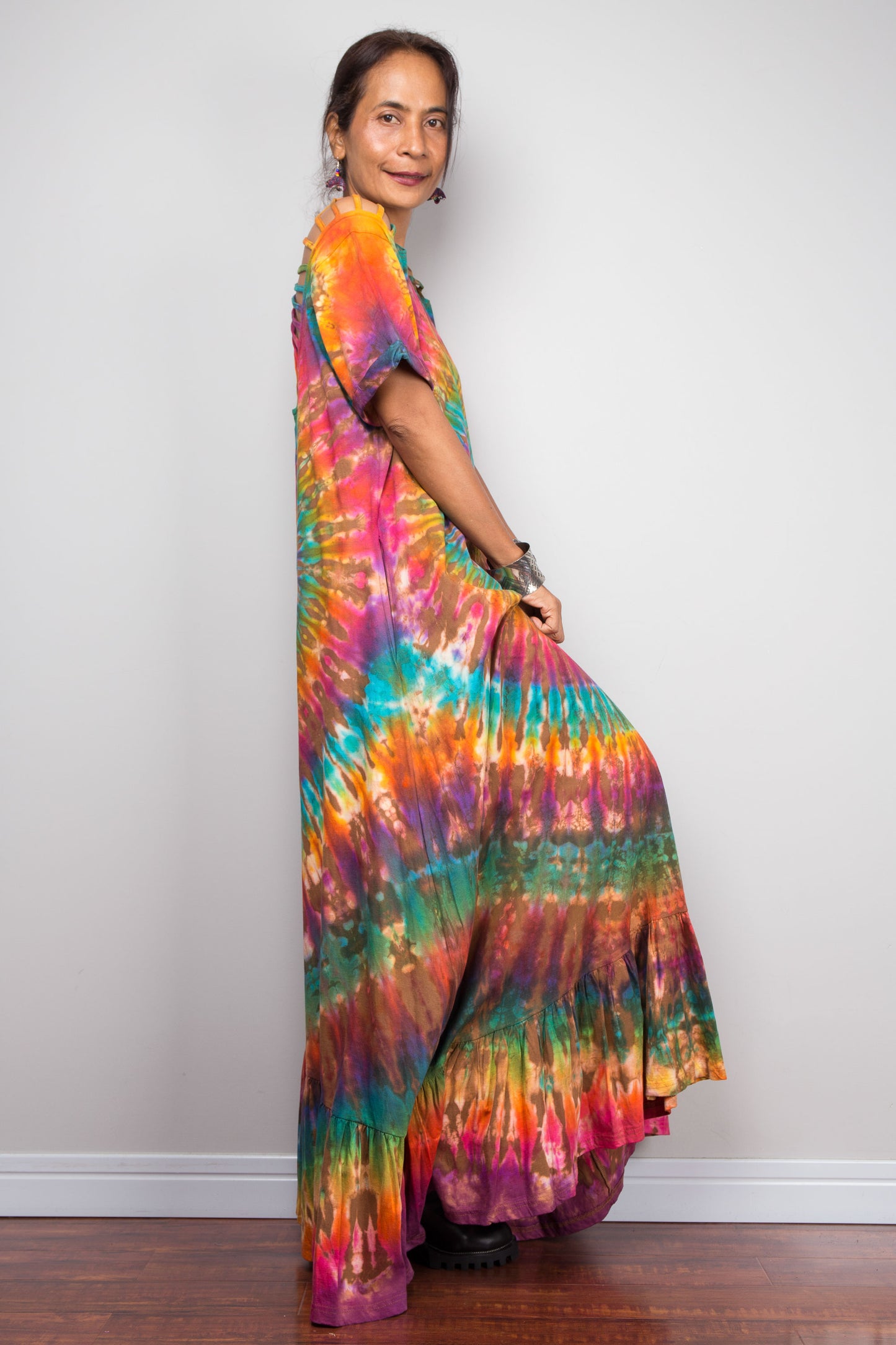 Side view of a petite model wearing the Nuichan rainbow tie dye maxi dress, showcasing the full-spectrum starburst pattern radiating from a central teal diamond, scoop neckline, short sleeves with cut-out ladder slash details, and tiered ruffle hem.