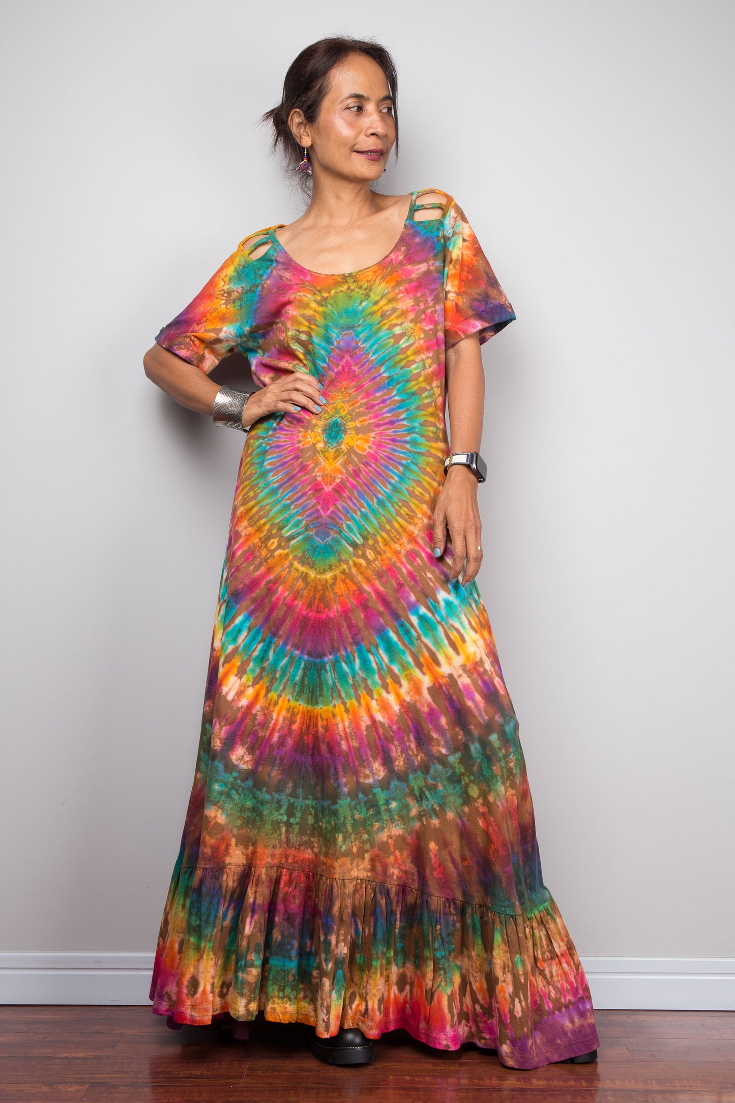 Front-facing view of a petite model wearing the Nuichan rainbow tie dye maxi dress with hand on one hip, showcasing the full-spectrum starburst pattern radiating from a central teal diamond, scoop neckline, short sleeves with cut-out ladder slash details, and tiered ruffle hem.