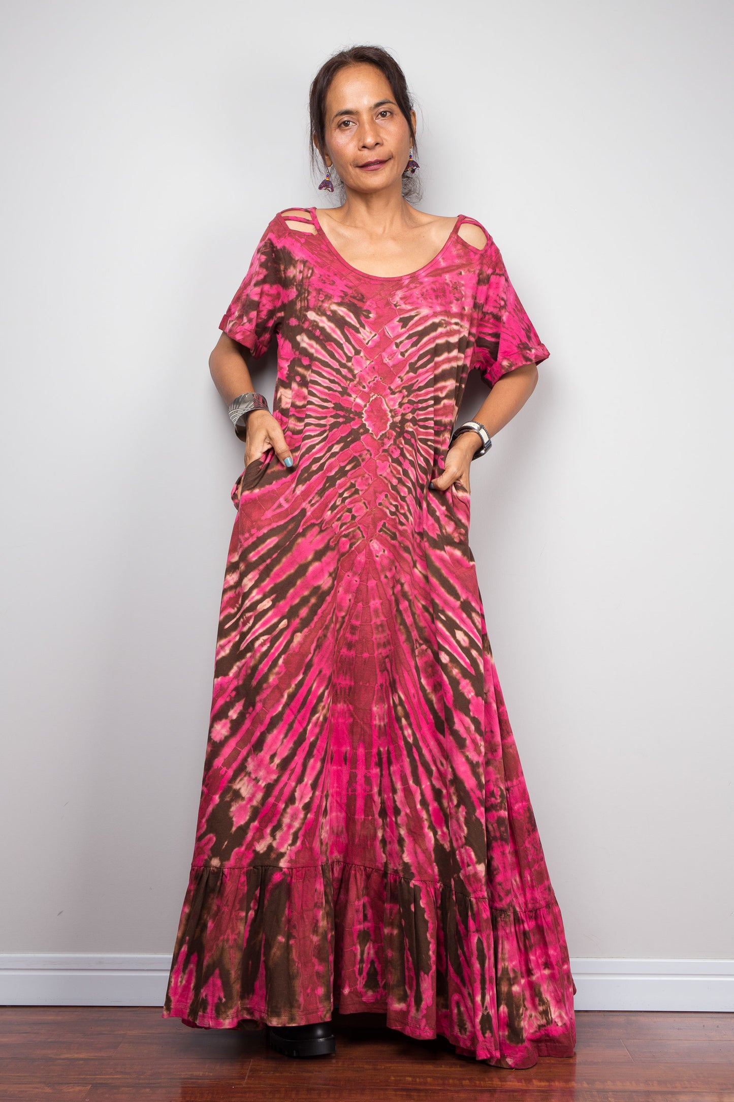 Front-facing view of a model wearing the Nuichan pink and brown tie dye maxi dress with hands in side pockets, showcasing the bold hot pink and chocolate brown starburst pattern, scoop neckline, short slashed sleeves.
