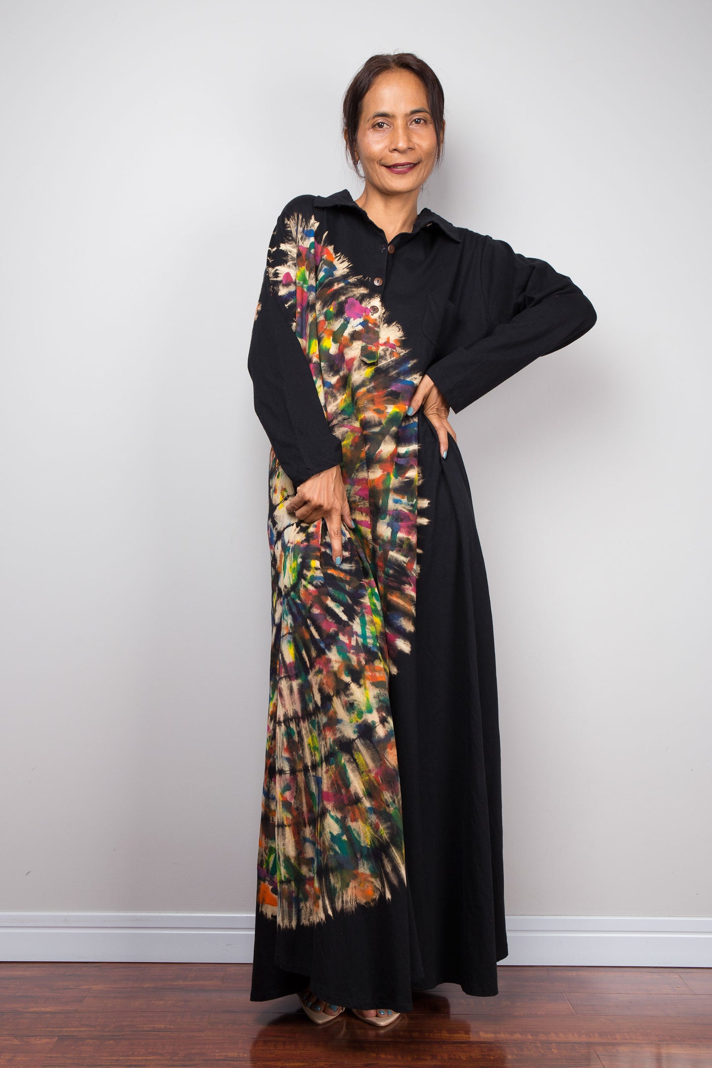 Petite woman in black long-sleeve maxi shirt dress. An asymmetric, vibrant hand-dyed multi-color tie-dye sunburst sweeps diagonally across the front. 